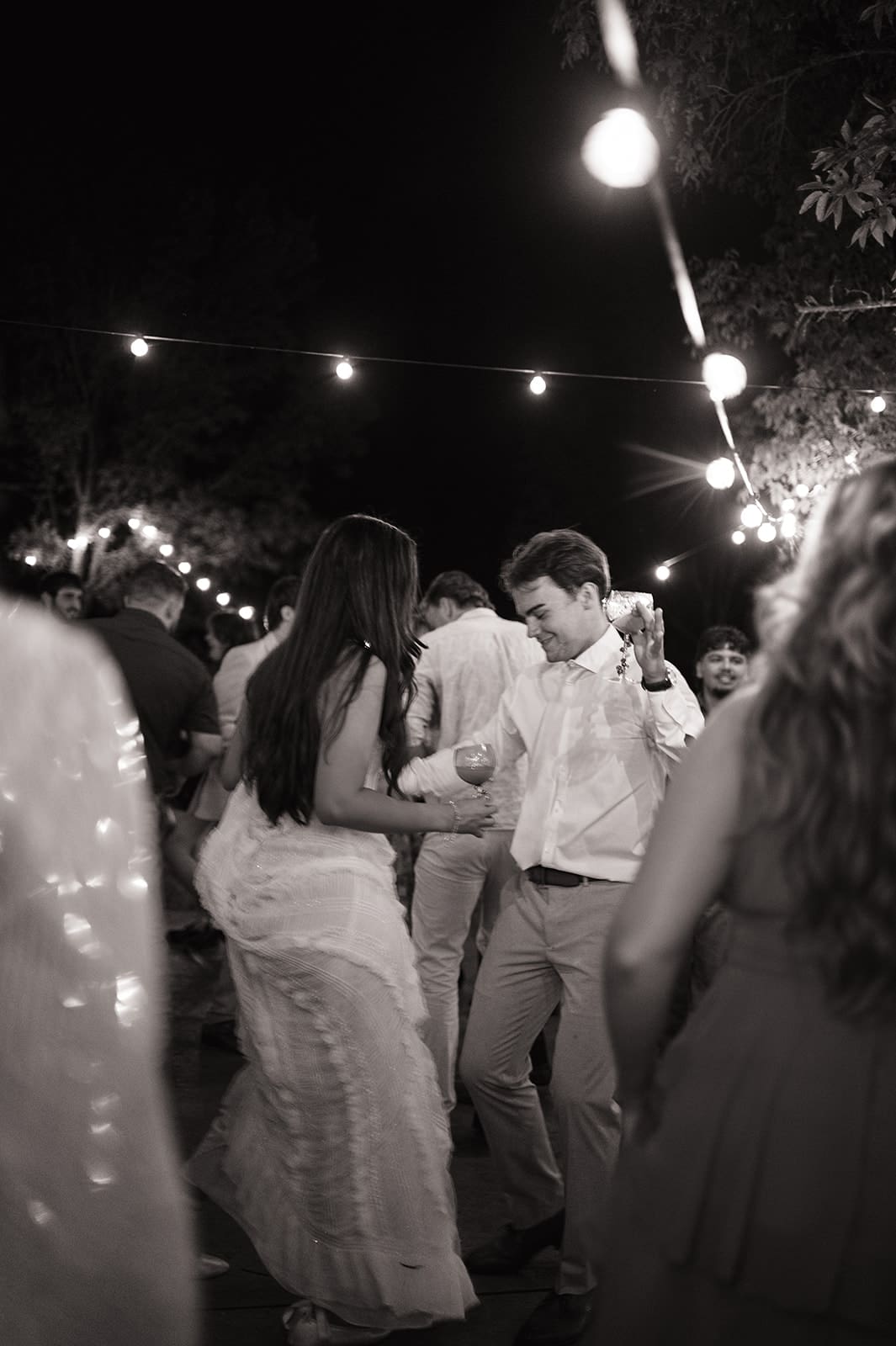 A bride and groom dance together at an outdoor night wedding reception, surrounded by guests and string lights for a backyard wedding