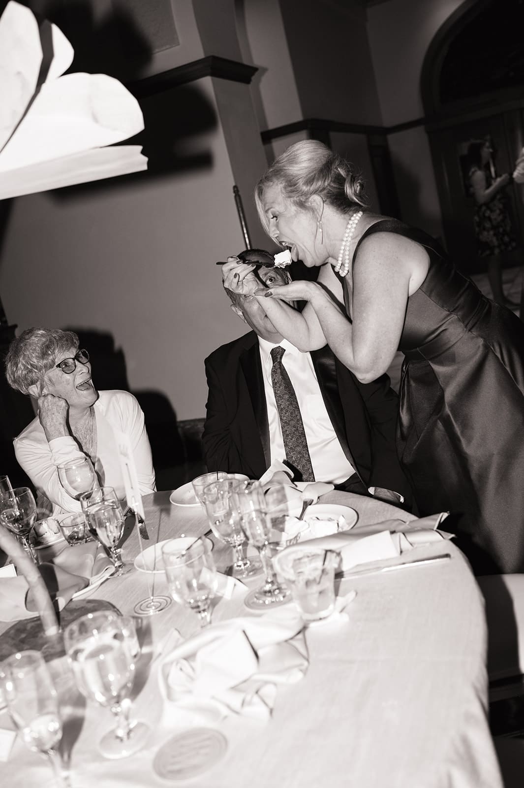 A woman in a dress stands and eats from a spoon at a formal dining table with two seated guests, surrounded by glasses and plates.