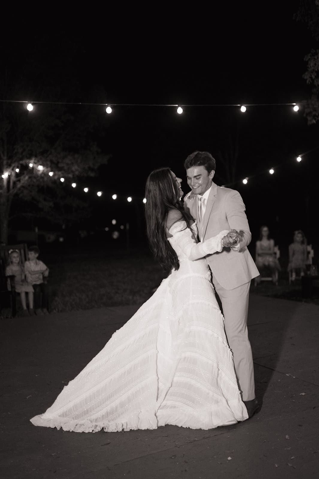 A bride in a white dress and a groom in a beige suit dance together outdoors at night under string lights for a backyard wedding in Texas