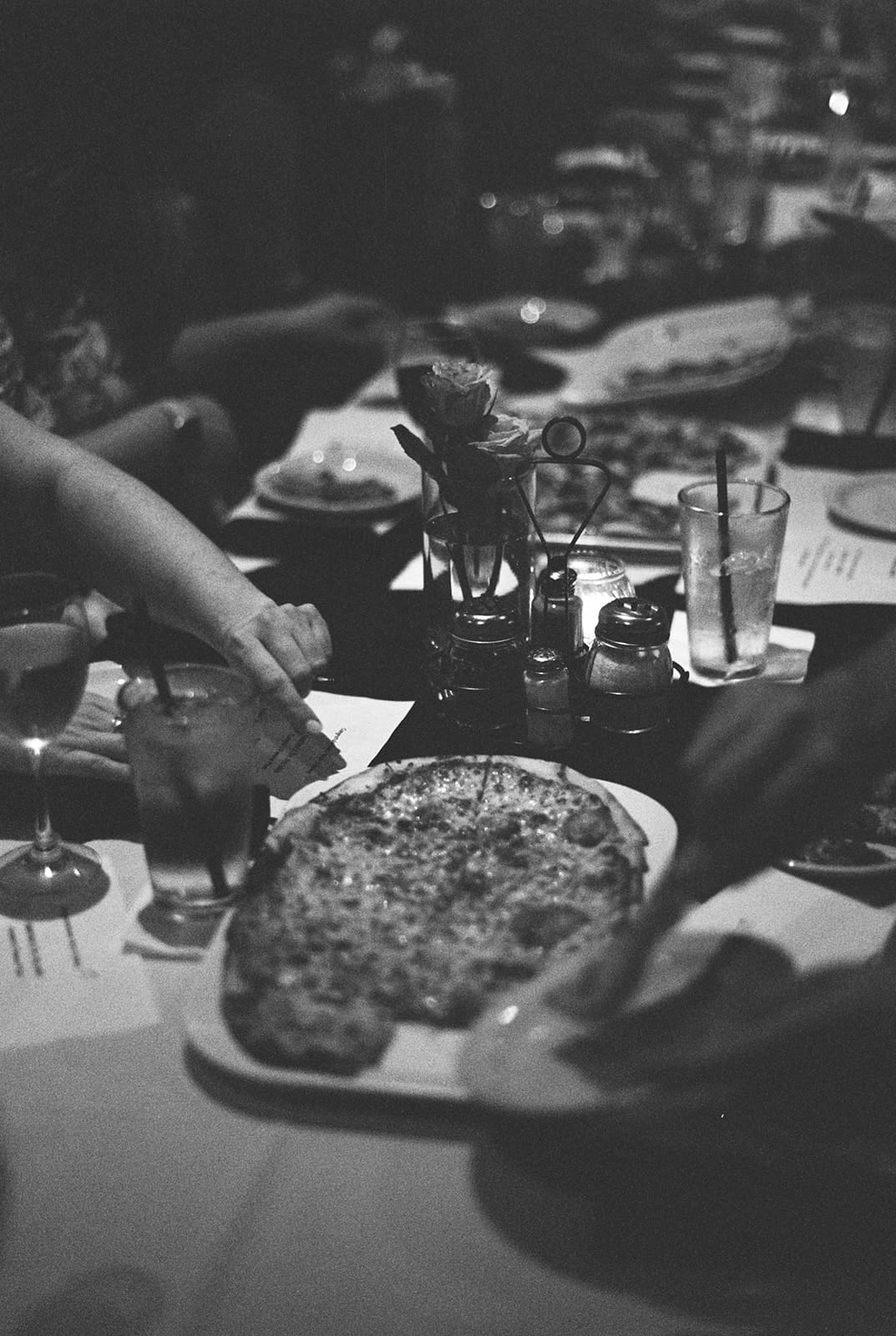 A group of people sit around a table with drinks and a large pizza, reaching for slices in a dimly lit restaurant.