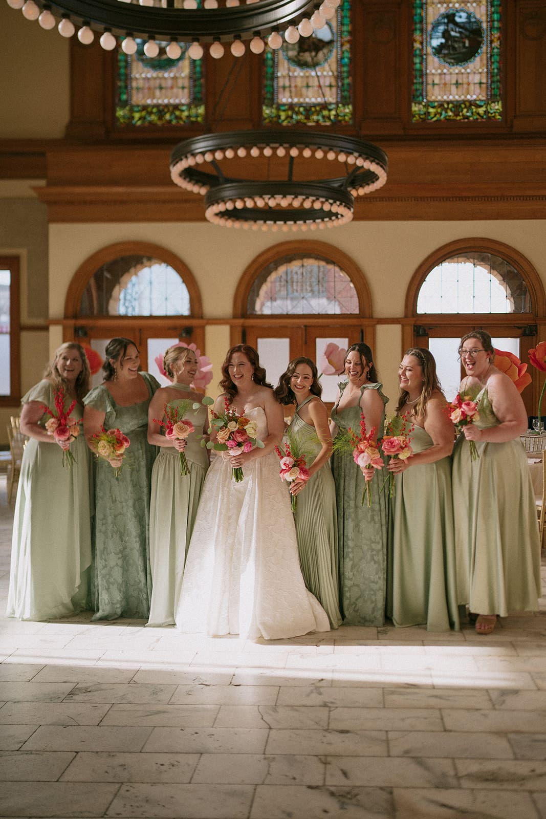 A bride in a white gown stands surrounded by seven bridesmaids in sage green dresses, all holding bouquets, inside a large, elegant hall with chandeliers at The Ashton Depot in Fort Worth
