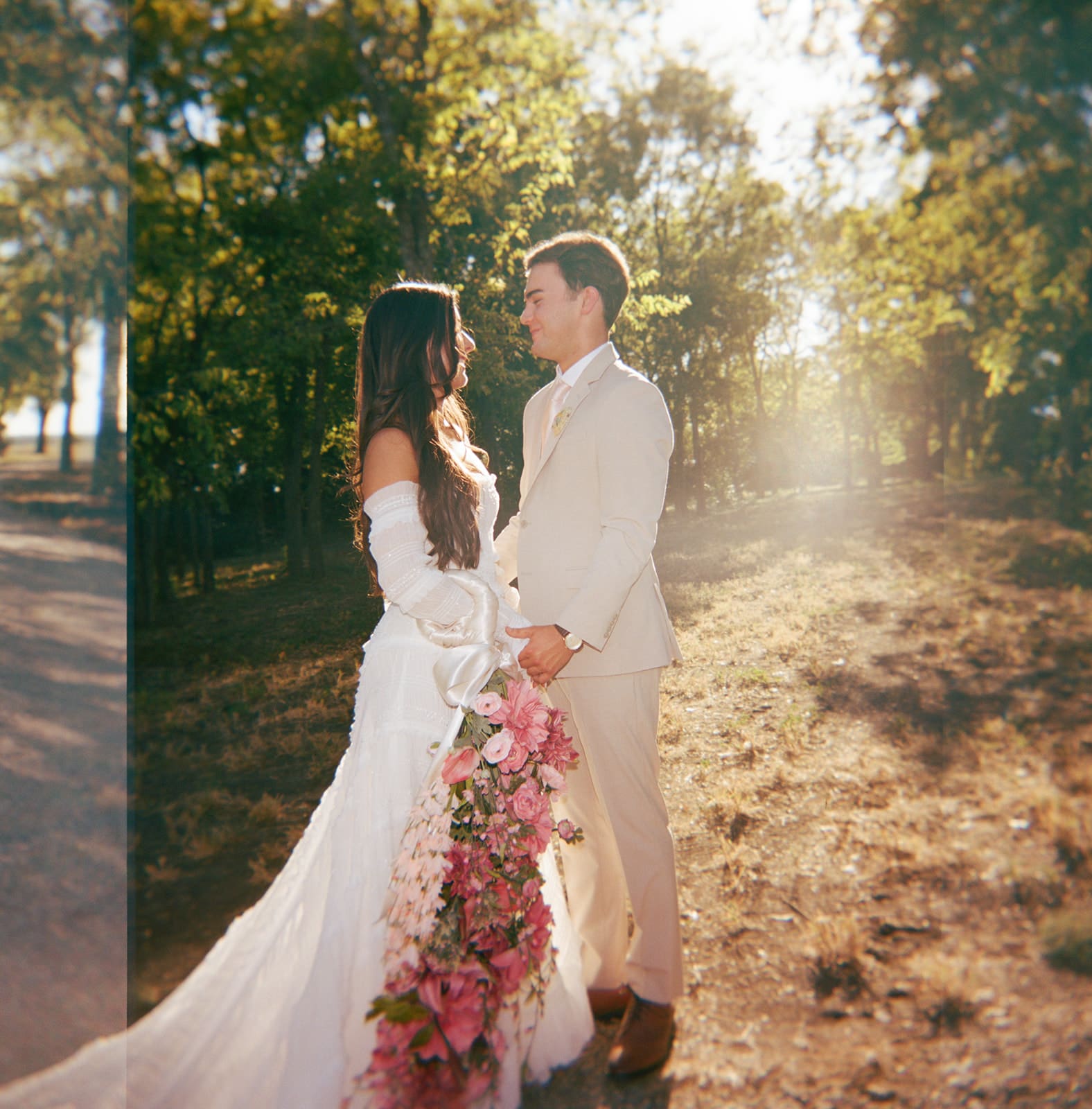 A bride and groom stand outdoors facing each other, holding hands in sunlight, with the bride holding a large bouquet of pink flowers for a backyard wedding in texas