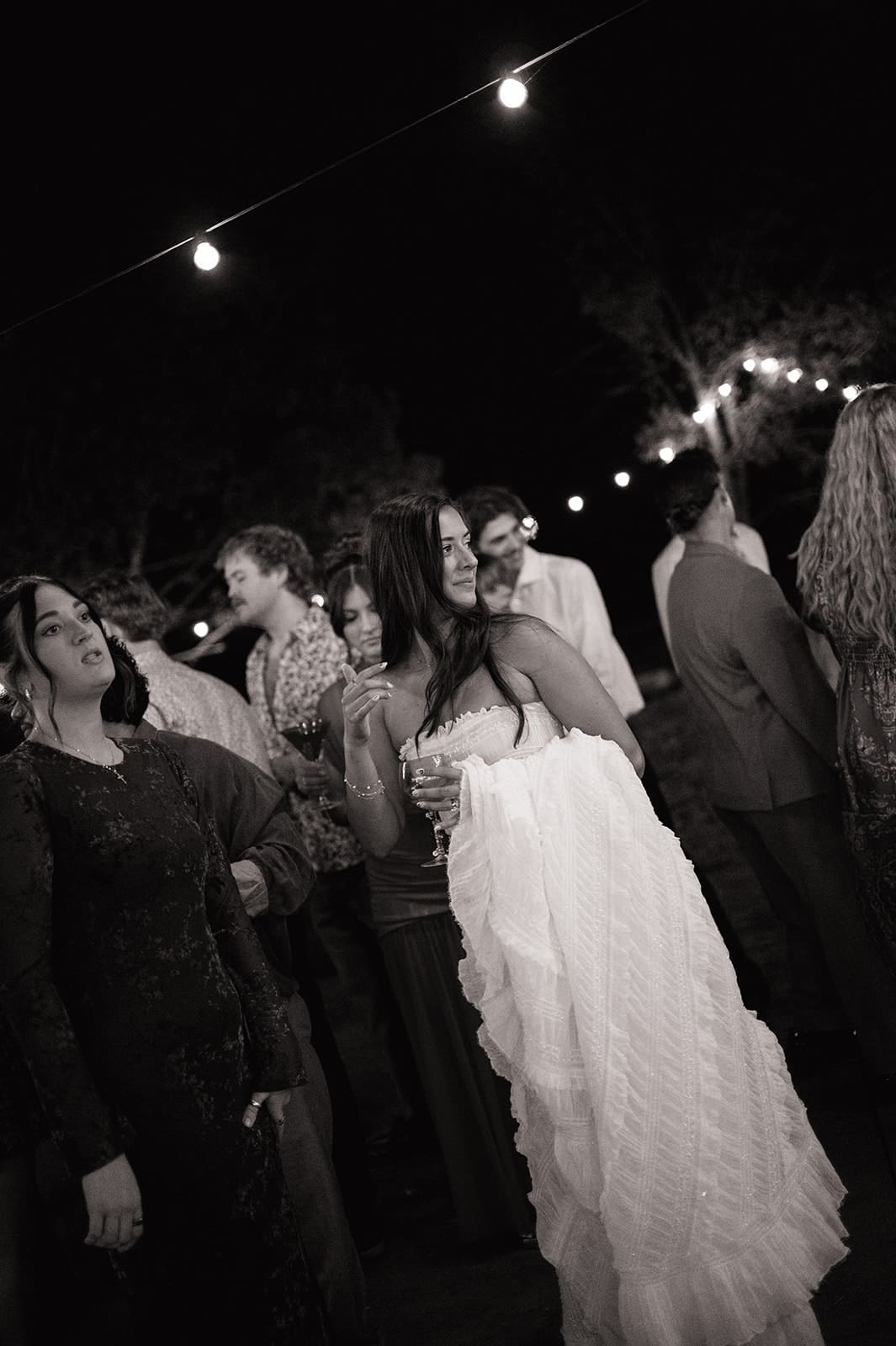A bride and groom dance together at an outdoor night wedding reception, surrounded by guests and string lights for a backyard wedding