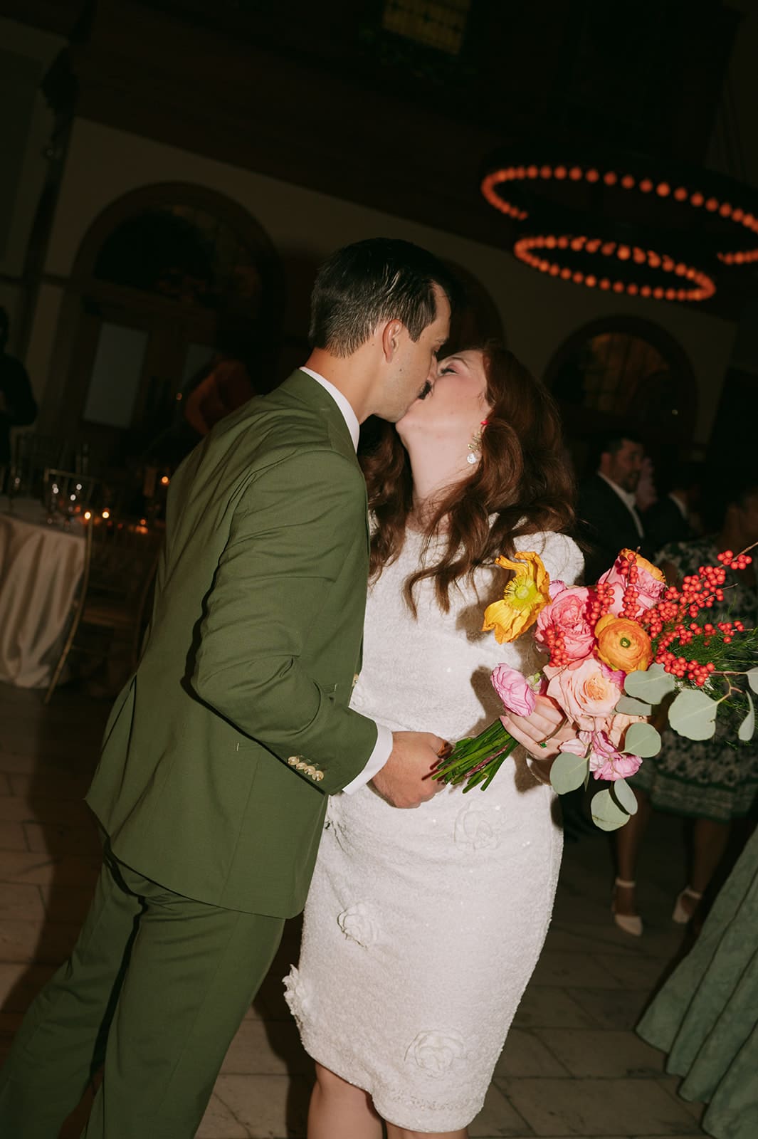 A man in a green suit and a woman in a white dress holding a bouquet sit together on a tiled floor, smiling at the camera for a wedding at The Ashton Depot in Fort Worth