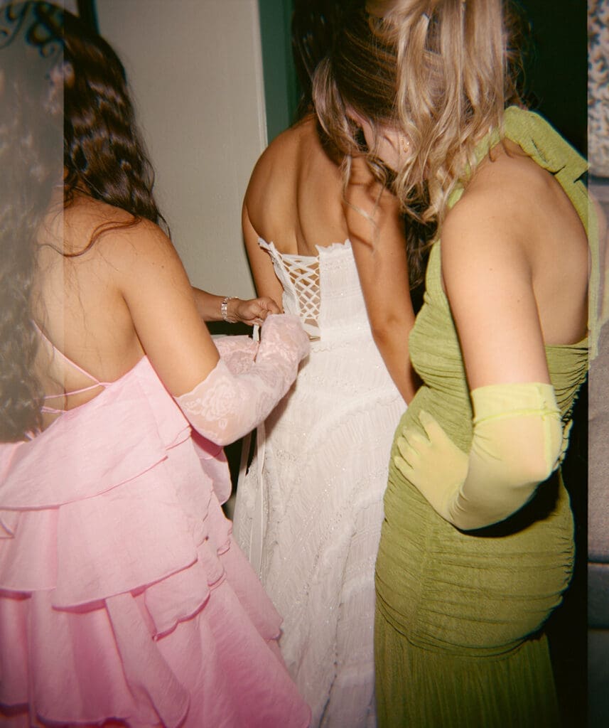 Three women in formal dresses stand together; one in pink helps another in white with the back of her dress, while the third, in a green dress and gloves, stands nearby.