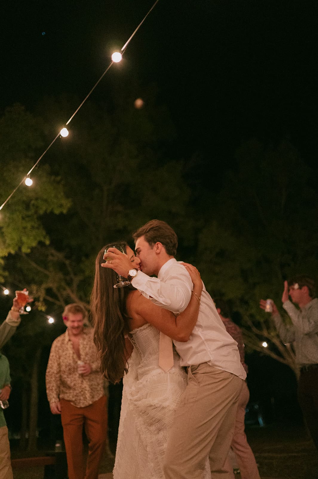 A bride and groom dance together at an outdoor night wedding reception, surrounded by guests and string lights for a backyard wedding
