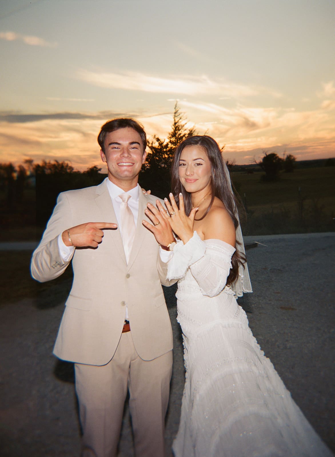 A couple in formal attire stands outdoors at sunset. The woman shows off a ring on her finger while the man smiles and points at her hand.