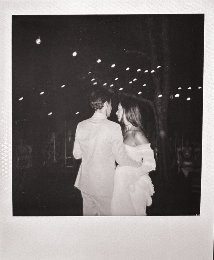 A couple dressed formally embraces and faces each other under string lights at night in a black and white photograph their backyard wedding