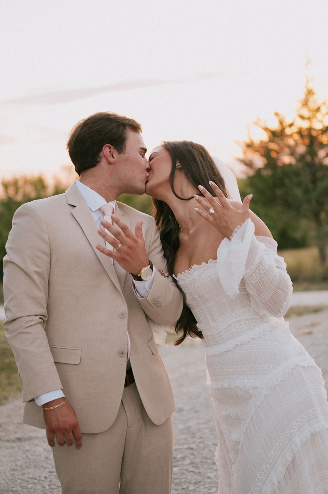 A bride and groom kiss outdoors at sunset; the bride shows her ring while wearing a white dress and the groom wears a beige suite at their backyard wedding 