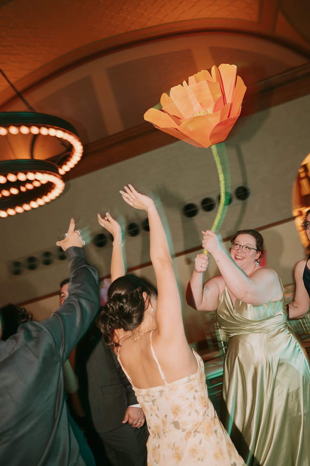 People in formal attire dance indoors; one person in a gold dress holds a large artificial orange flower above their head.