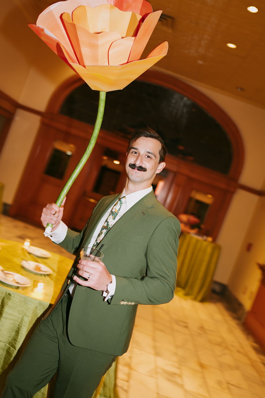 People in formal attire dance indoors; one person in a gold dress holds a large artificial orange flower above their head.