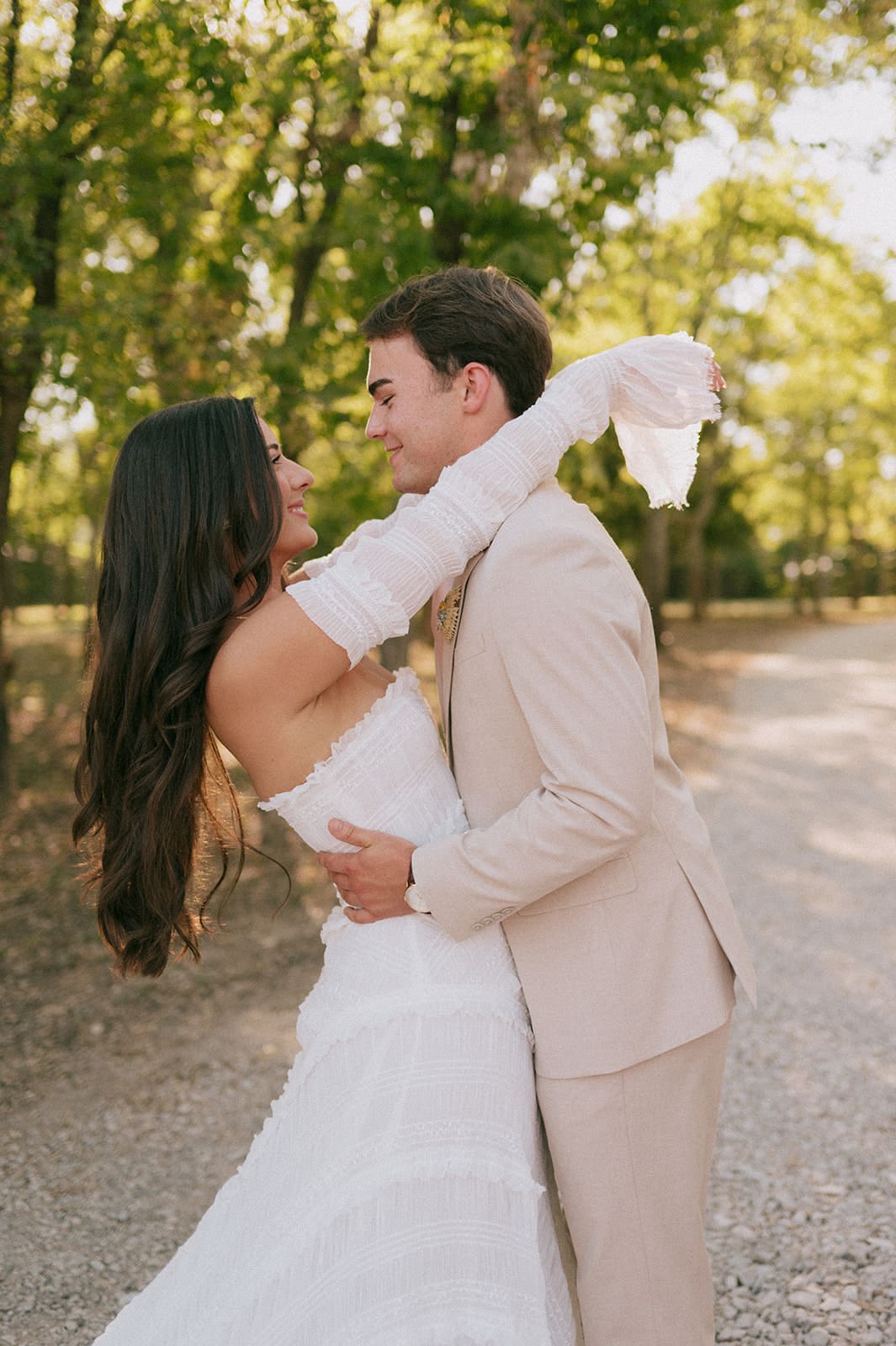 A bride and groom stand outdoors facing each other, holding hands in sunlight, with the bride holding a large bouquet of pink flowers for a backyard wedding in texas