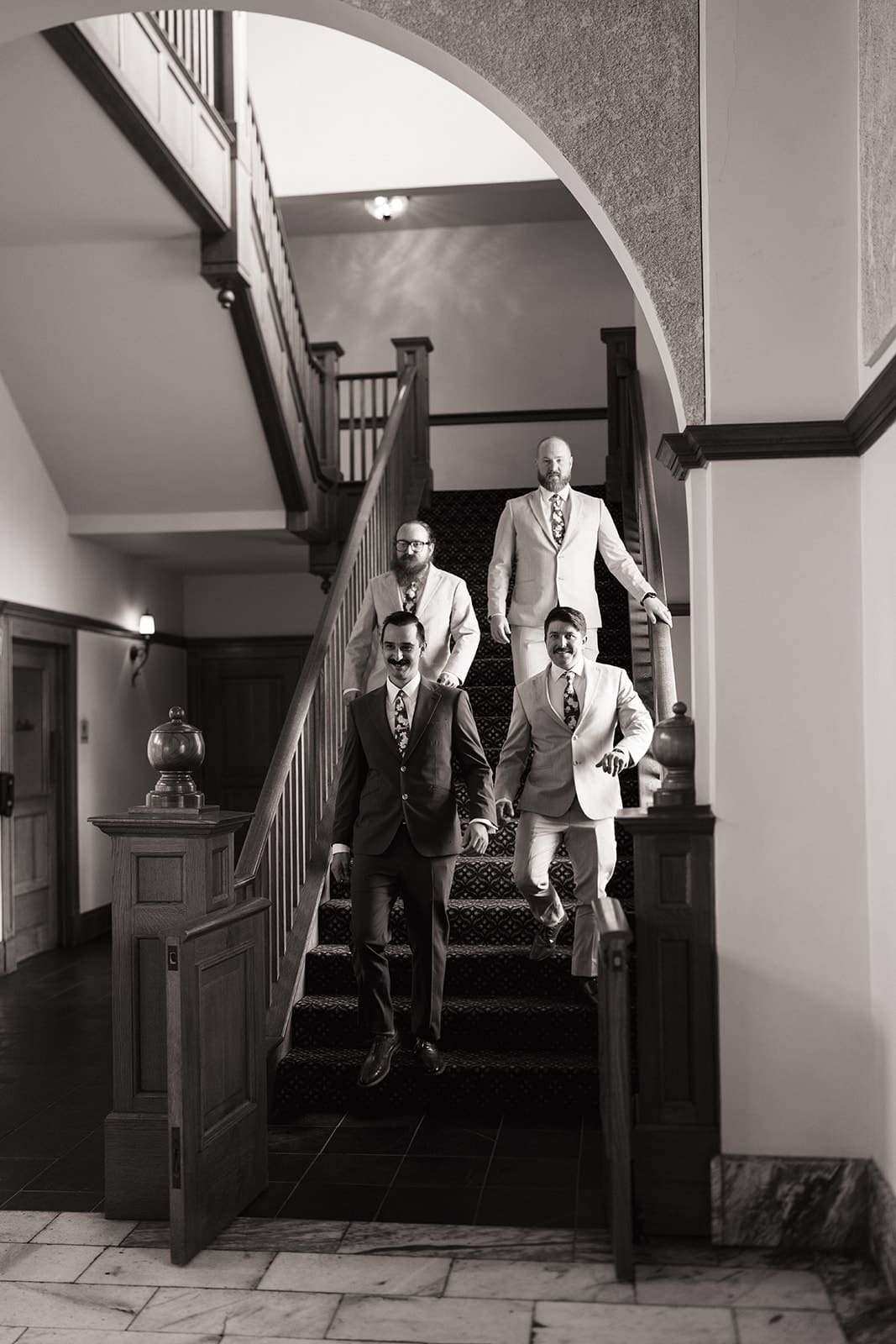 Four men in suits descend a staircase in a formal indoor setting. The photo is in black and white for a edding at The Ashton Depot in Fort Worth