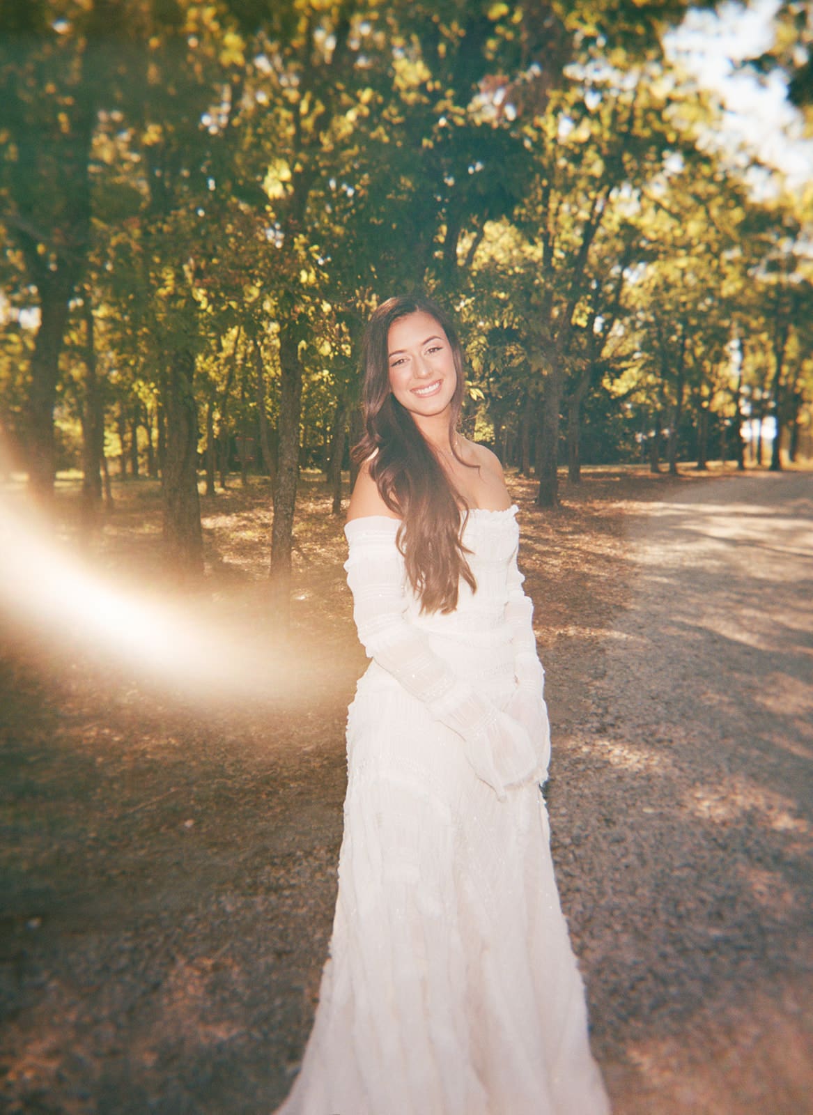 A woman in a white wedding dress stands outdoors on a tree-lined path, smiling at the camera.