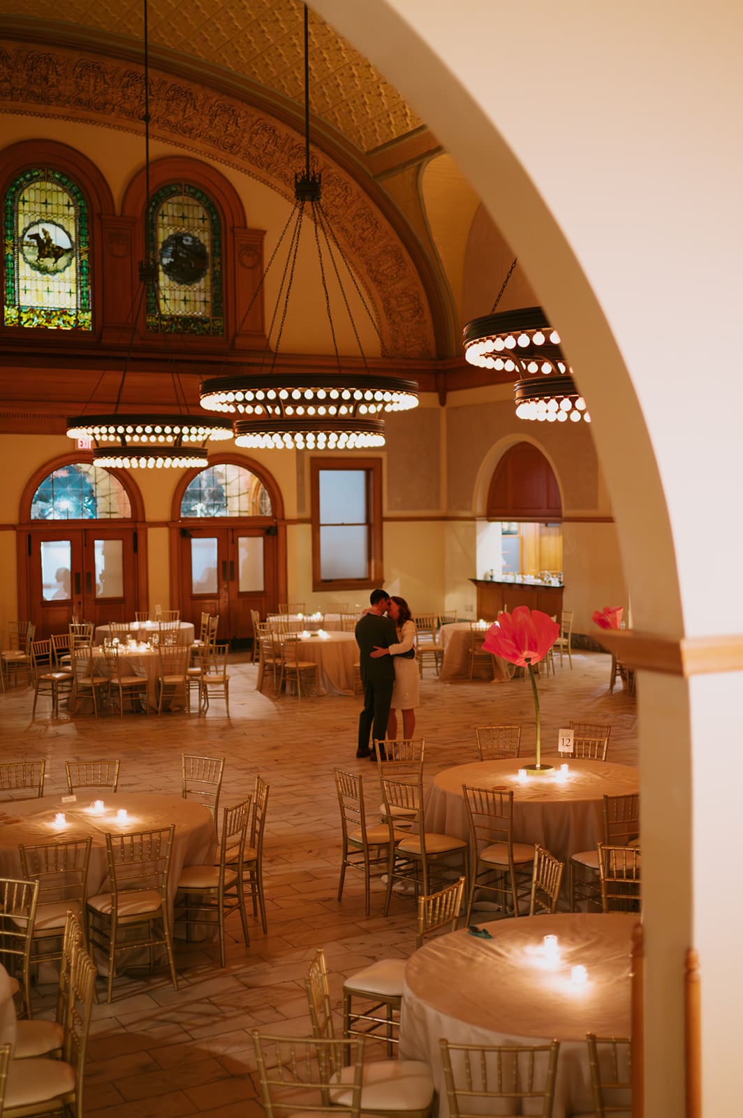 A couple stands together in the center of a large, elegant hall with round tables, wooden chairs, and stained glass windows.