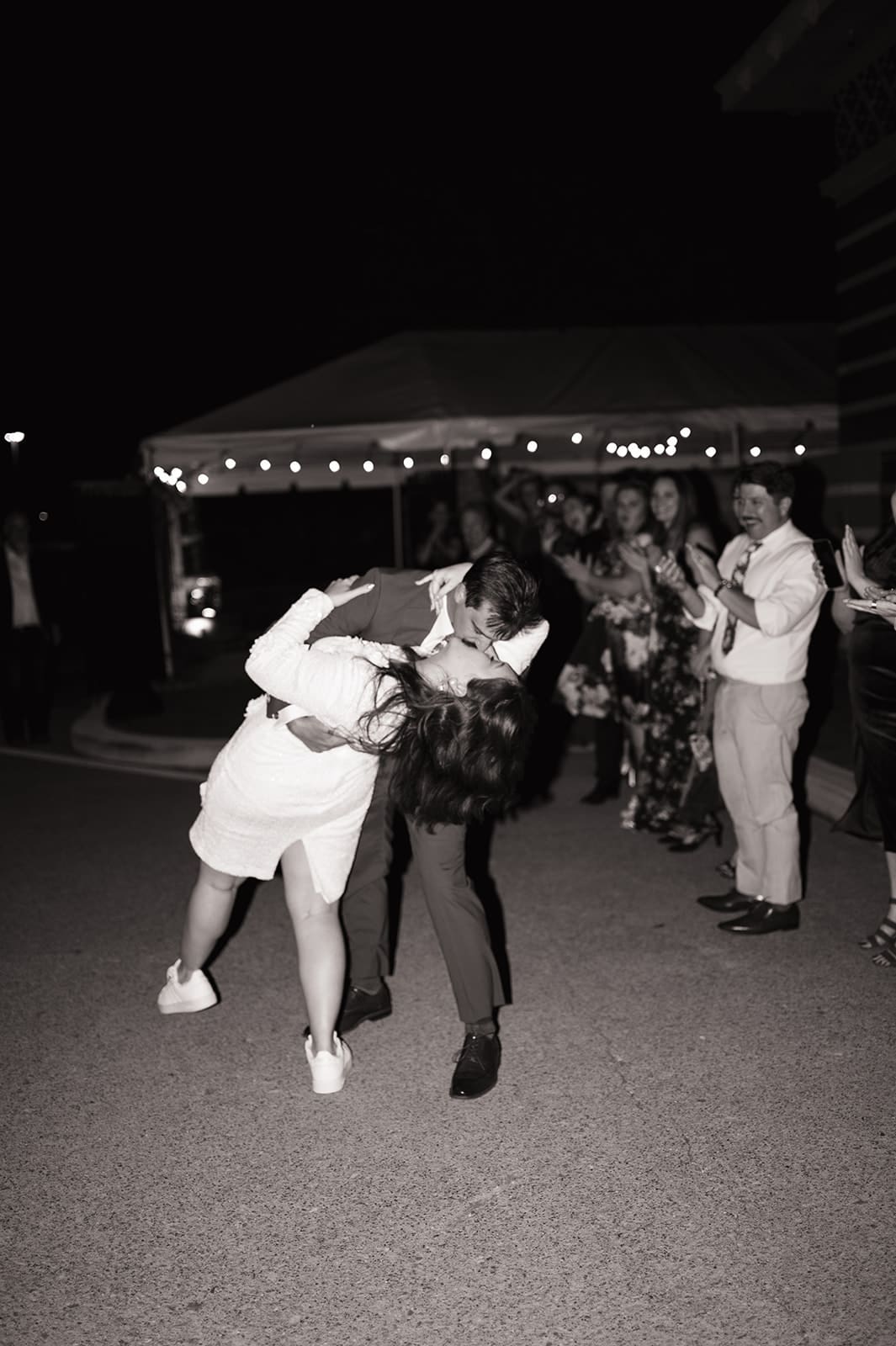 A couple dances under string lights at night; the man dips the woman while people in the background watch and clap.