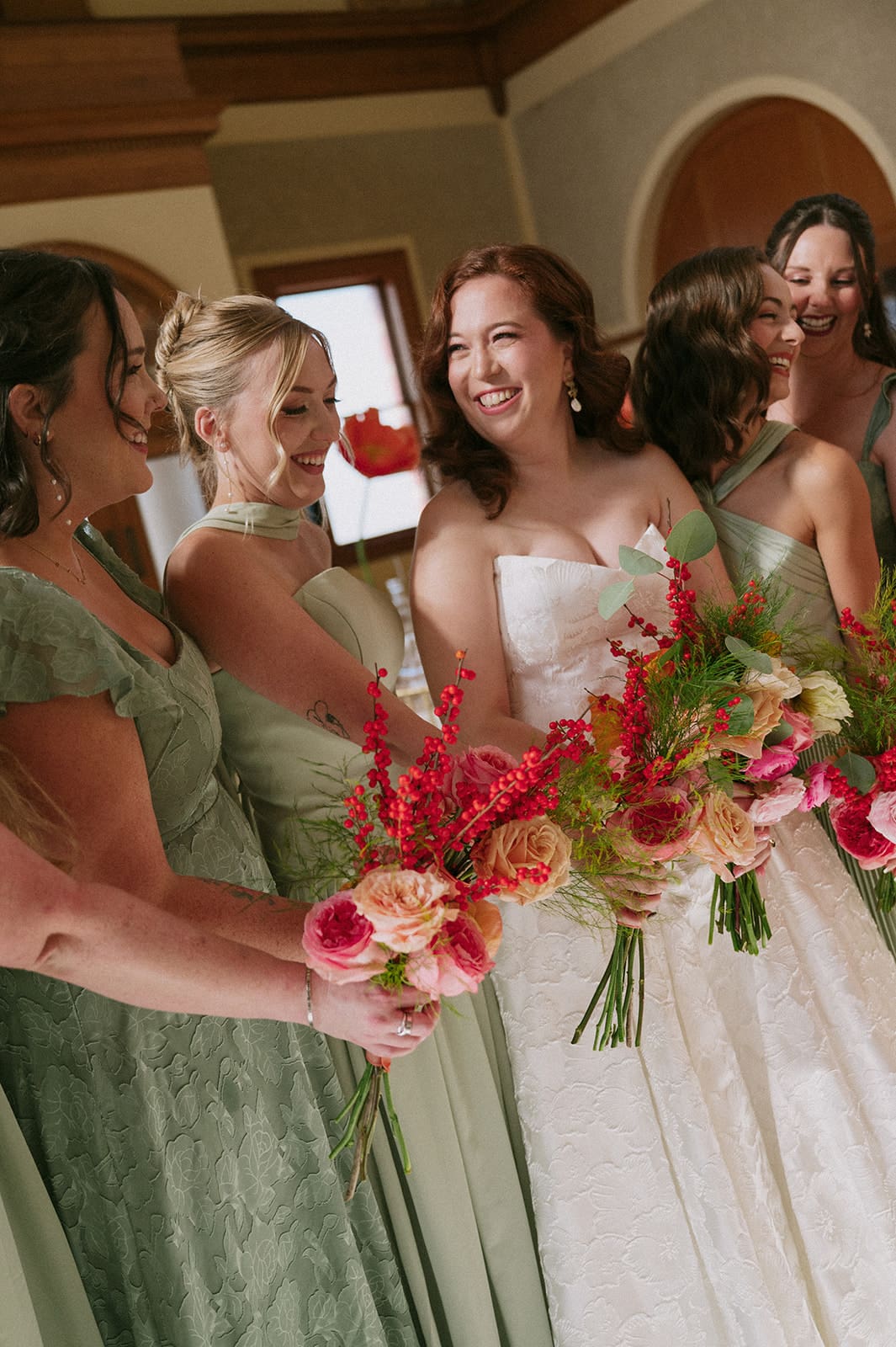 A bride in a white gown stands surrounded by seven bridesmaids in sage green dresses, all holding bouquets, inside a large, elegant hall with chandeliers at The Ashton Depot in Fort Worth