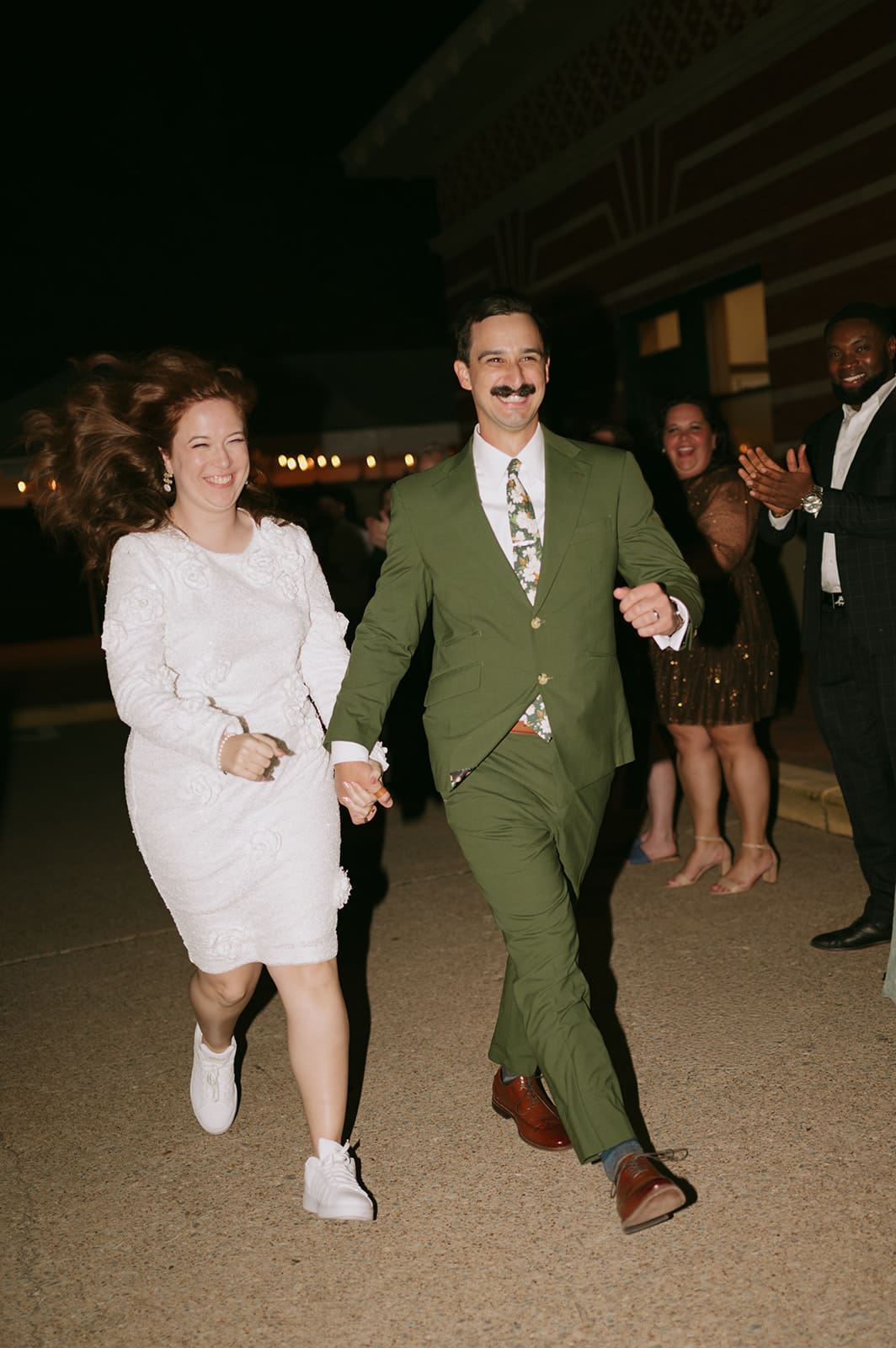 A smiling couple, the woman in a white dress and sneakers and the man in a green suit, hold hands and run while people clap in the background at night.