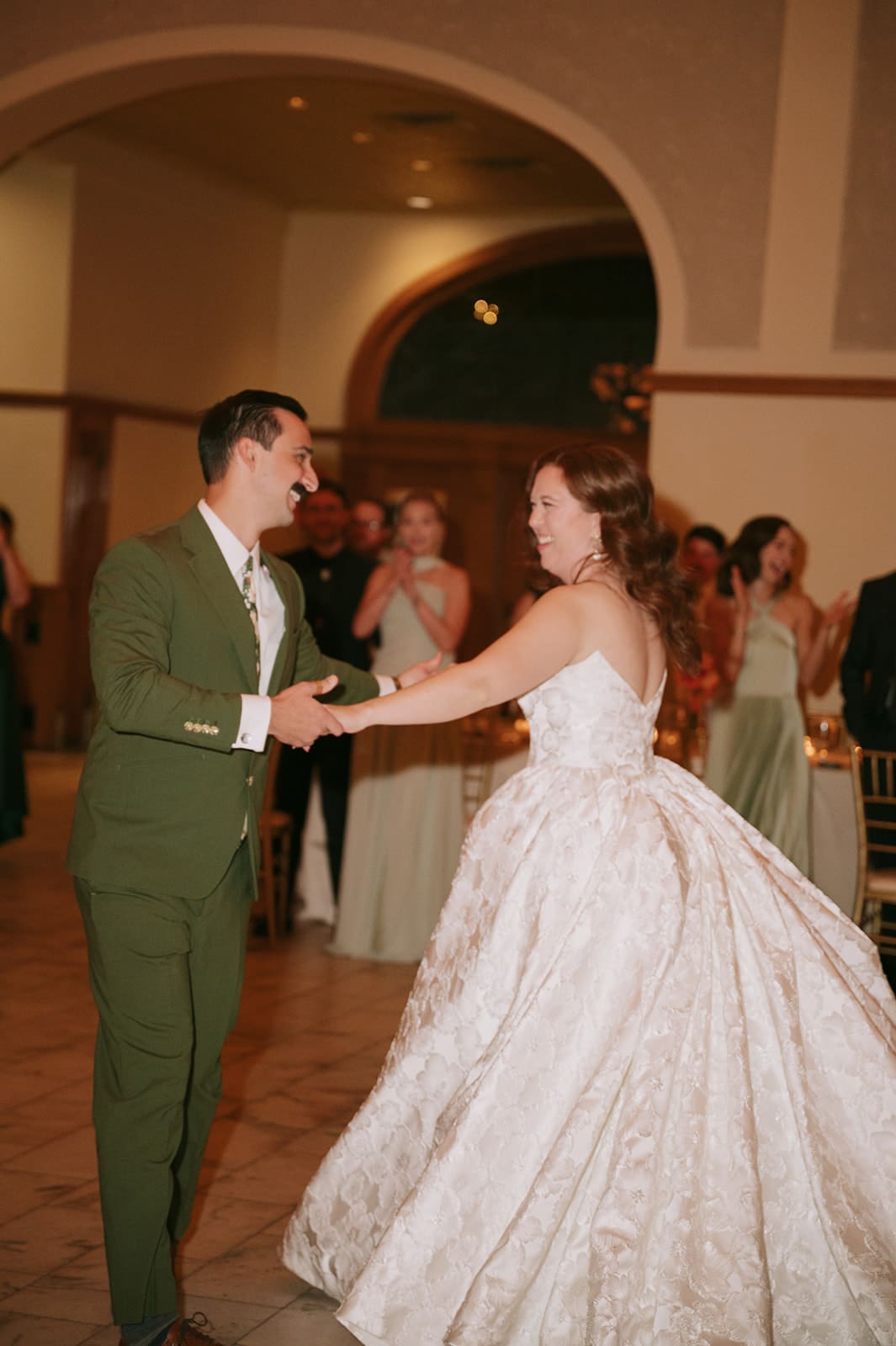 A couple dances together at a formal event; the woman wears a white gown and the man wears a green suit. Other guests watch in the background.