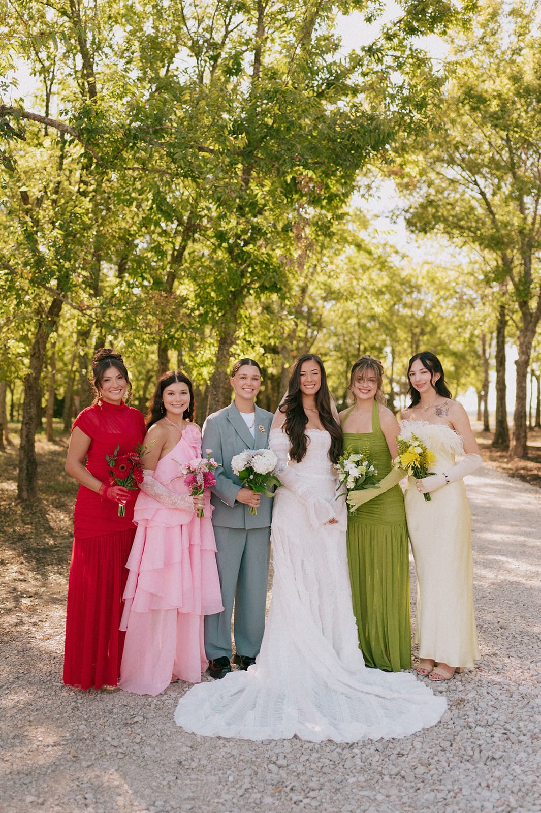 A group of six people, dressed in formal attire, stand together outdoors on a sunlit tree-lined path, smiling at the camera for a backyard wedding in Texas