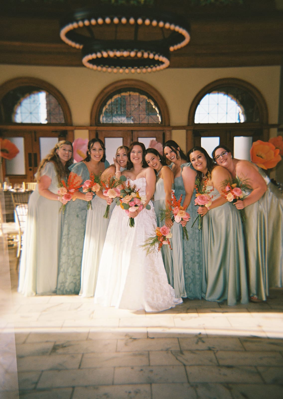 A bride in a white gown stands surrounded by seven bridesmaids in sage green dresses, all holding bouquets, inside a large, elegant hall with chandeliers at The Ashton Depot in Fort Worth