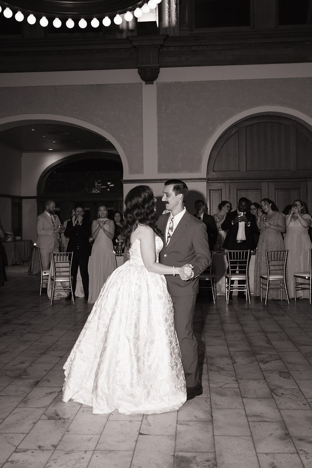 A couple dances together at a formal event; the woman wears a white gown and the man wears a green suit. Other guests watch in the background.