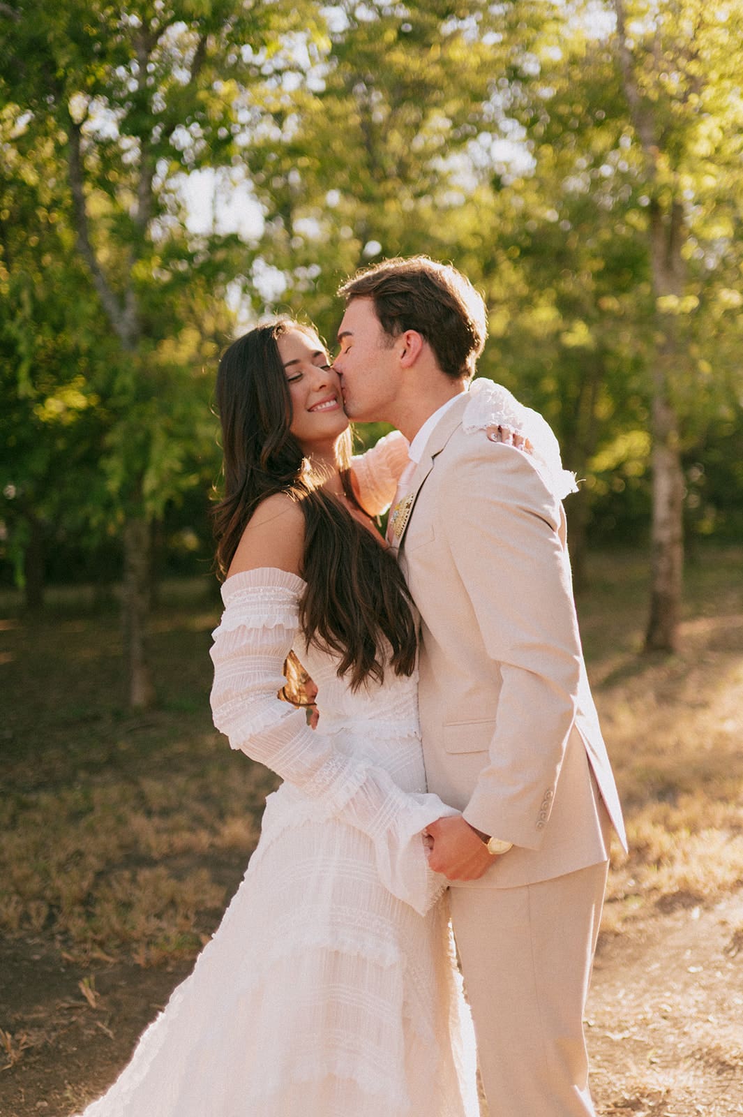 A bride and groom stand outdoors facing each other, holding hands in sunlight, with the bride holding a large bouquet of pink flowers for a backyard wedding in texas