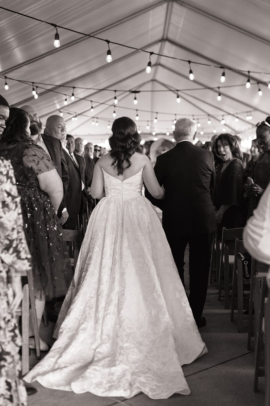A bride in a white gown holding a bouquet walks down the aisle arm-in-arm with an older man at an indoor wedding ceremony with seated guests for a Wedding at The Ashton Depot in Fort Worth,