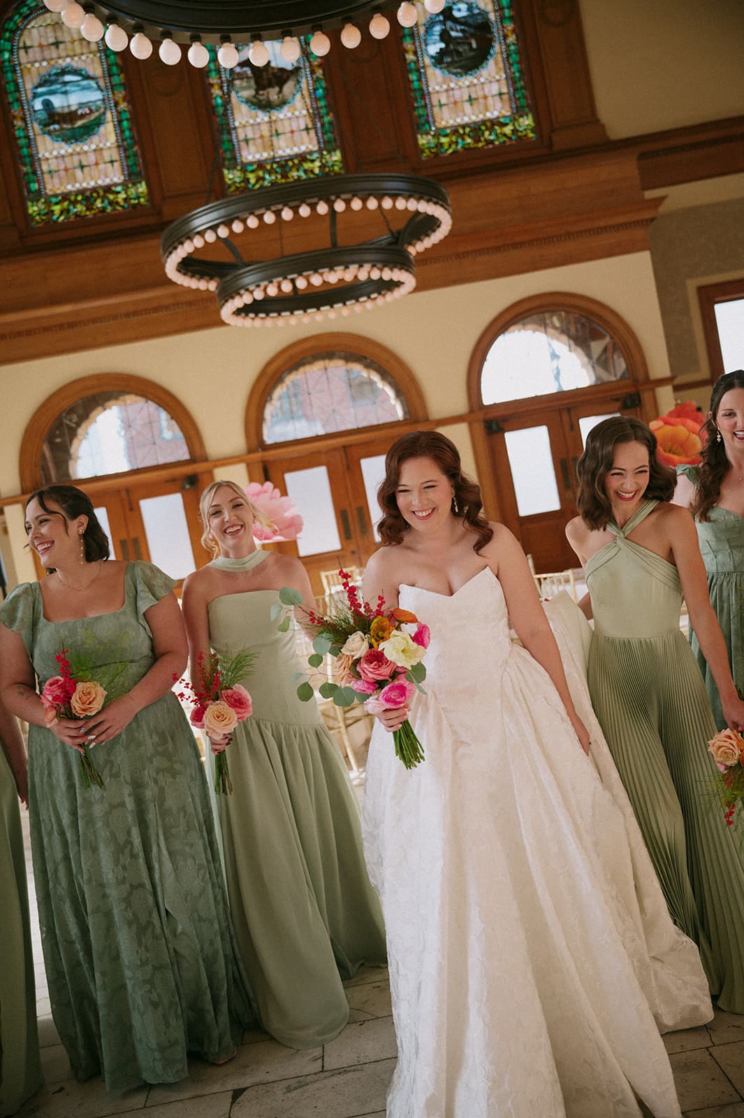 A bride in a white gown stands surrounded by seven bridesmaids in sage green dresses, all holding bouquets, inside a large, elegant hall with chandeliers at The Ashton Depot in Fort Worth