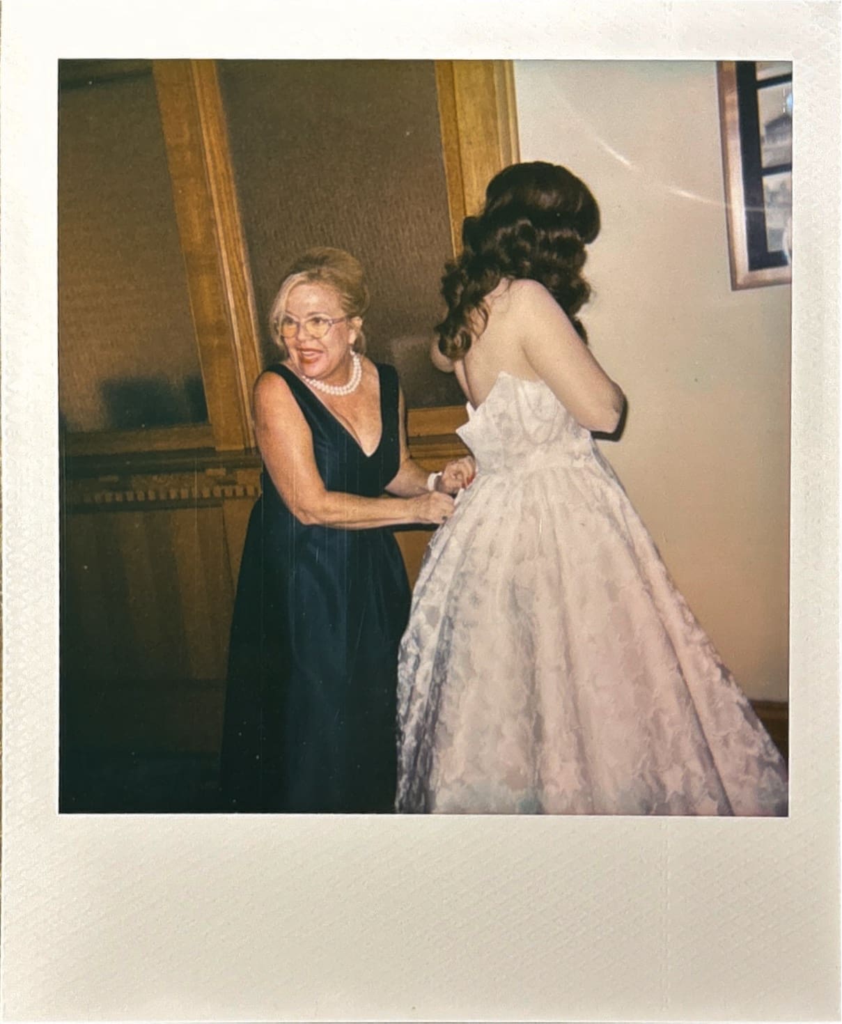 A woman in a dark dress helps another woman in a white gown adjust the back of her dress in an indoor setting for a Wedding at The Ashton Depot in Fort Worth