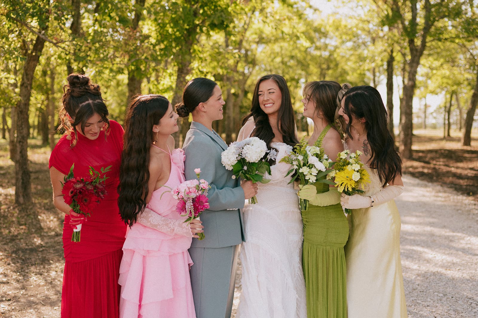 A group of six people, dressed in formal attire, stand together outdoors on a sunlit tree-lined path, smiling at the camera for a backyard wedding in Texas