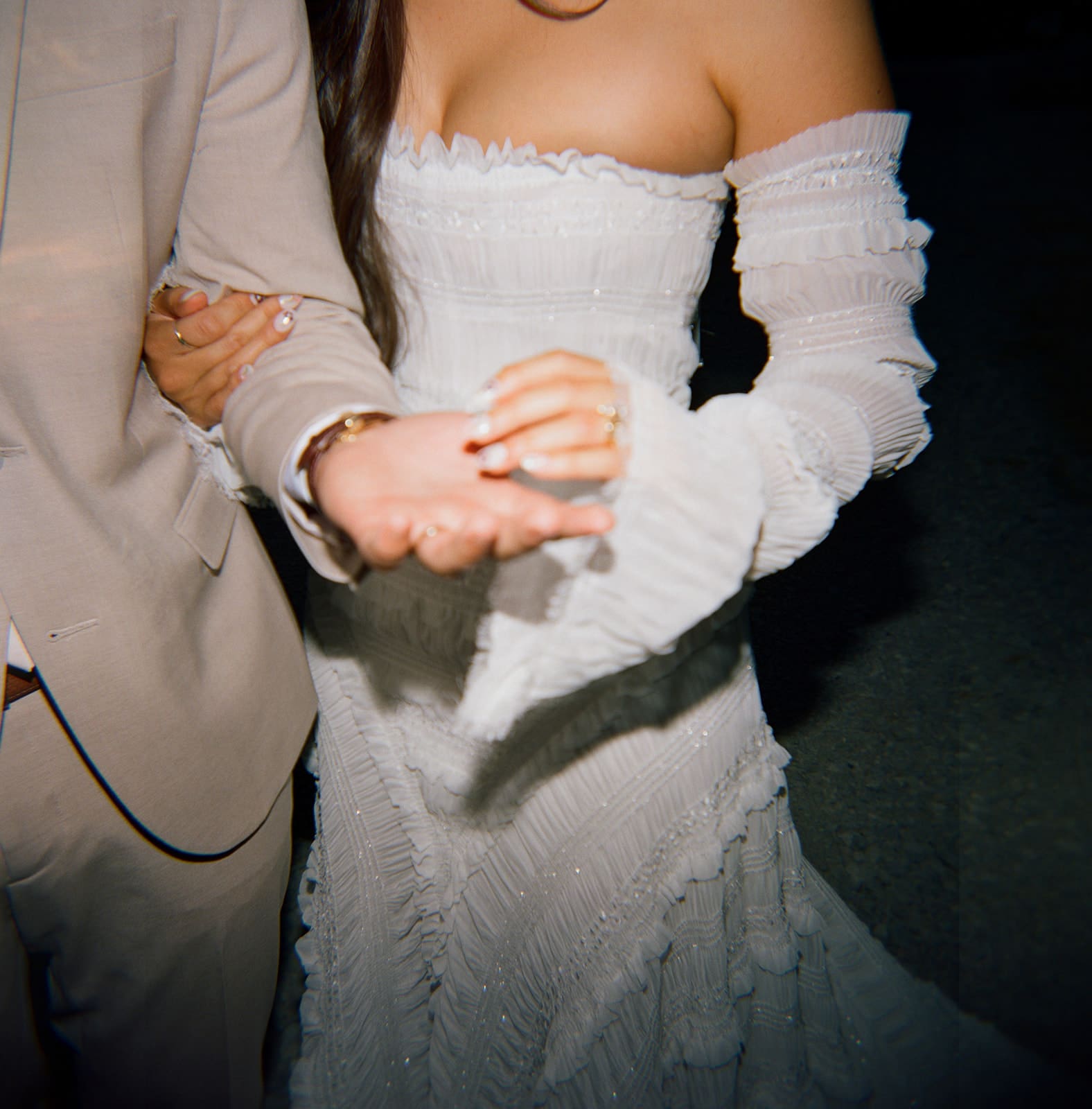 A couple in formal attire stands outdoors at sunset. The woman shows off a ring on her finger while the man smiles and points at her hand.