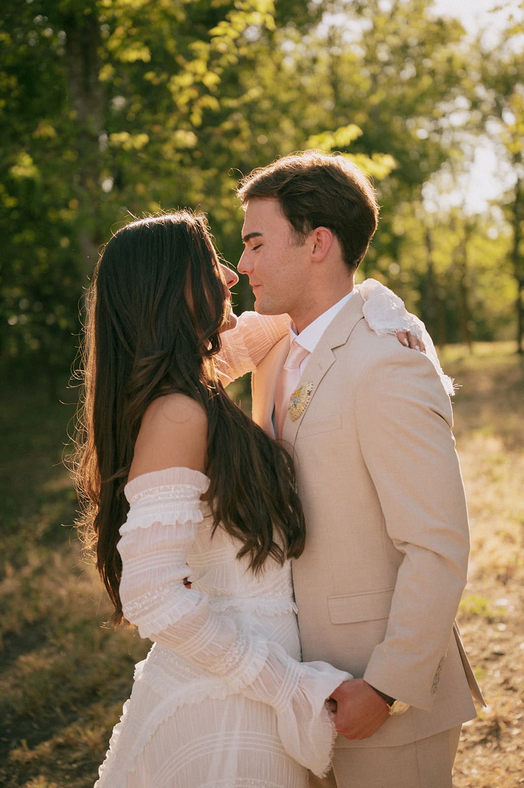 A bride and groom stand outdoors facing each other, holding hands in sunlight, with the bride holding a large bouquet of pink flowers for a backyard wedding in texas