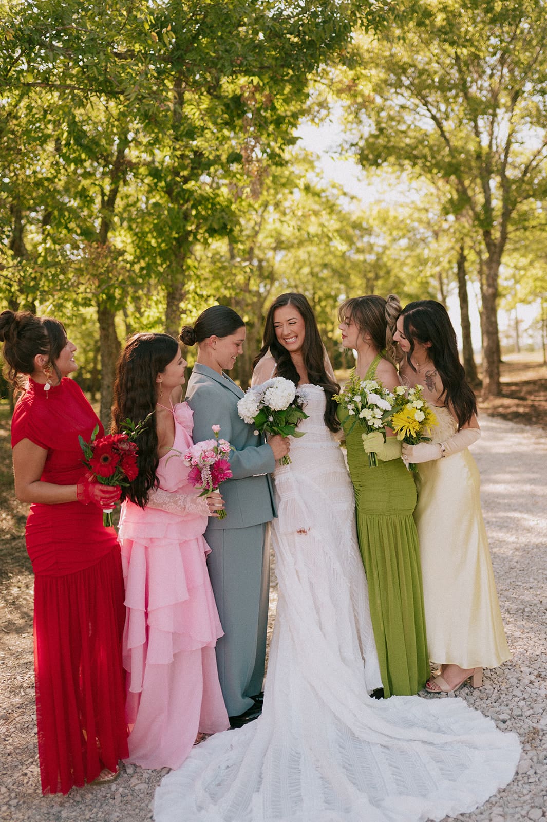 A group of six people, dressed in formal attire, stand together outdoors on a sunlit tree-lined path, smiling for a backyard wedding in Texas