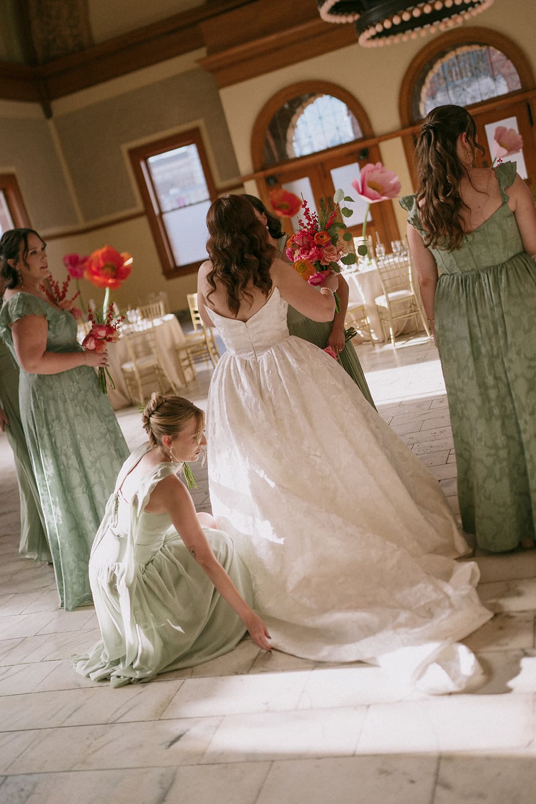 A bride in a white gown stands surrounded by seven bridesmaids in sage green dresses, all holding bouquets, inside a large, elegant hall with chandeliers at The Ashton Depot in Fort Worth