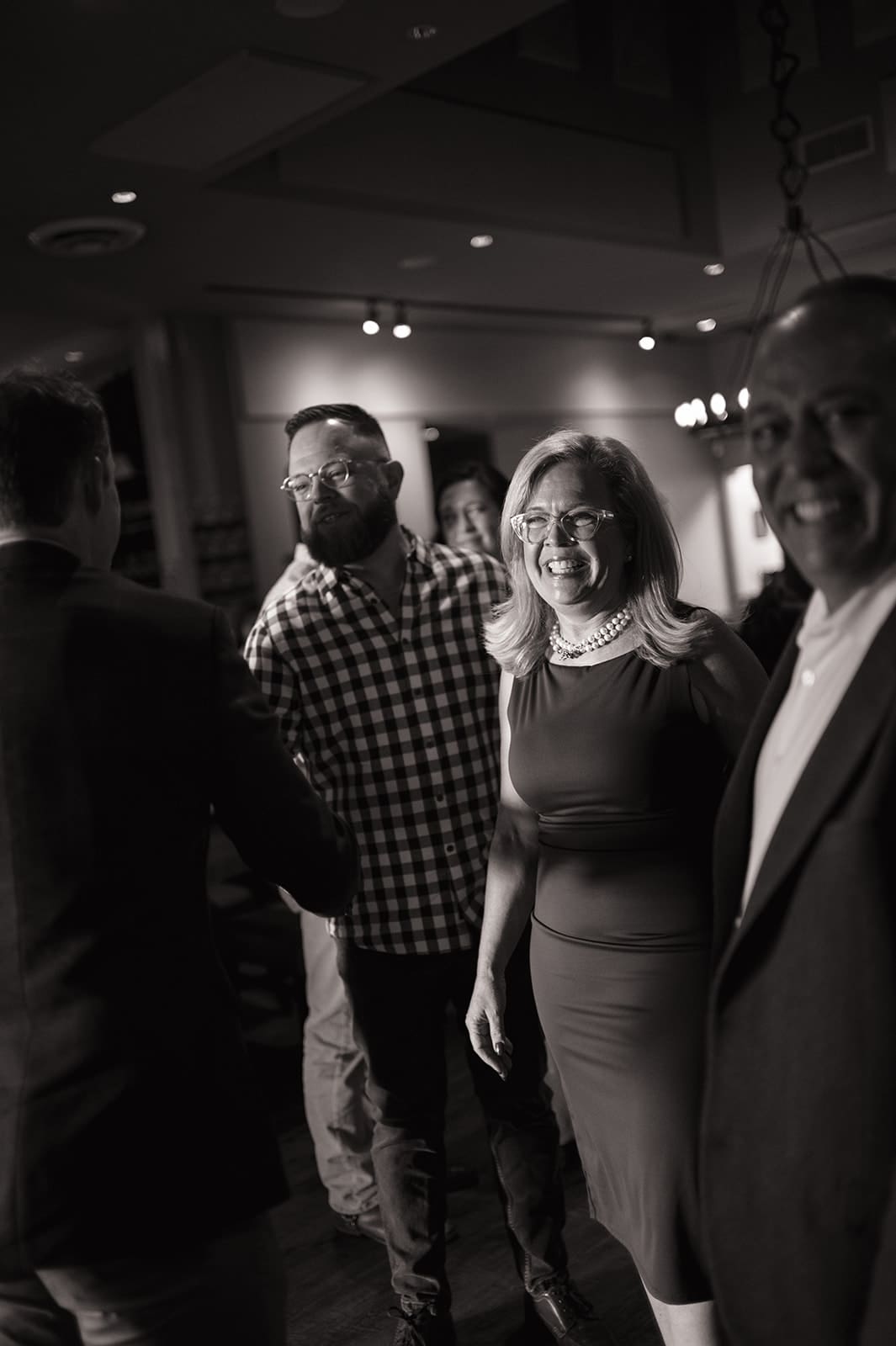 A group of four adults, dressed in semi-formal attire, stand together indoors and interact, smiling in a dimly lit room.