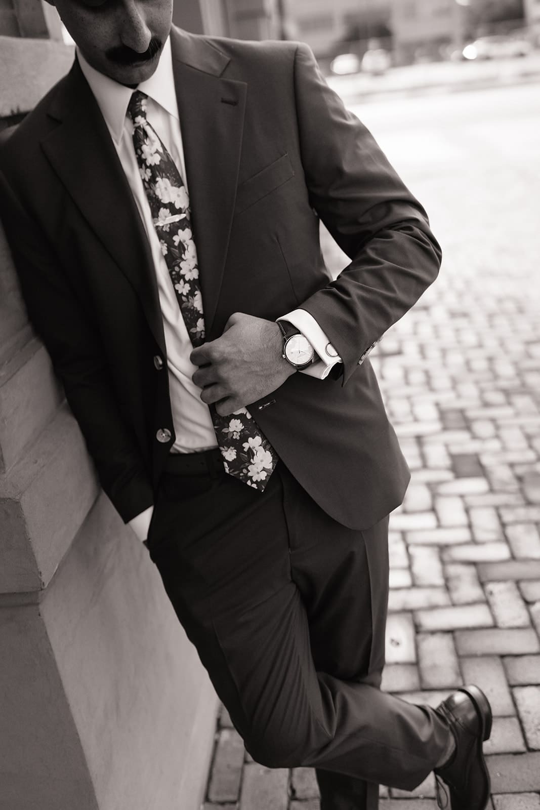 Man in a suit with a floral tie leans against a wall, looking down at his watch, standing on a brick sidewalk in an urban setting.