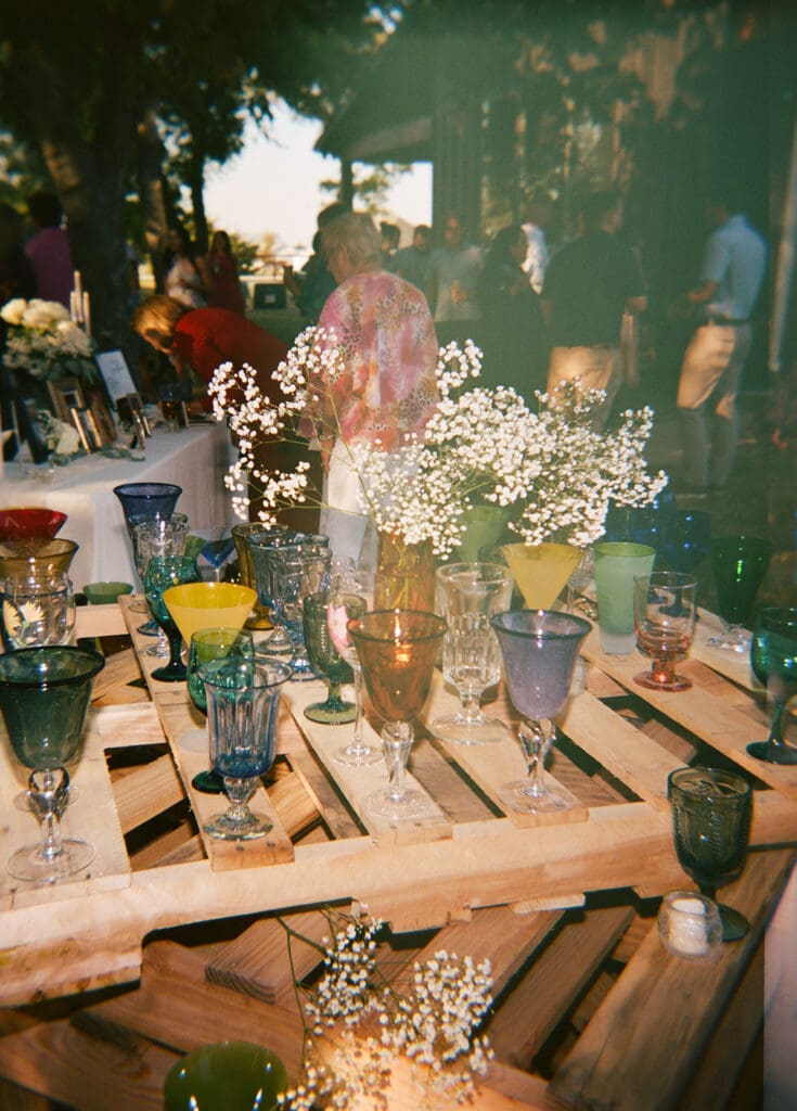 A variety of colorful glass goblets are arranged on a wooden pallet table, decorated with small white flowers, at an outdoor event.