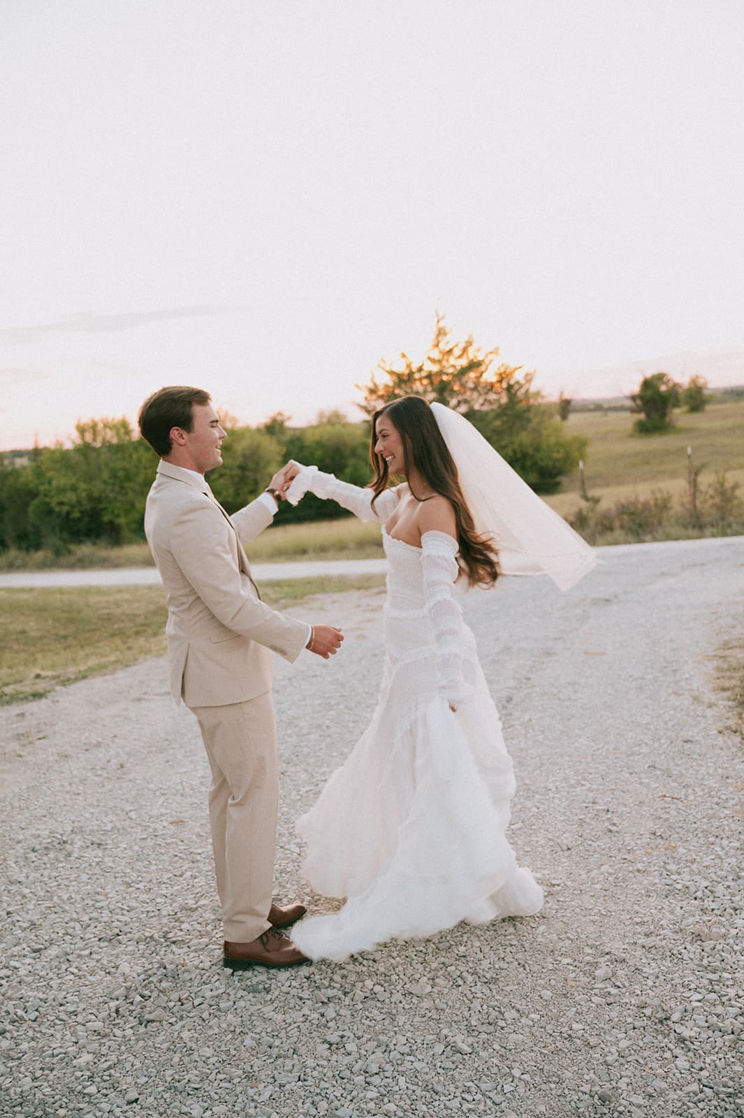 A bride and groom kiss outdoors at sunset; the bride shows her ring while wearing a white dress and the groom wears a beige suite at their backyard wedding