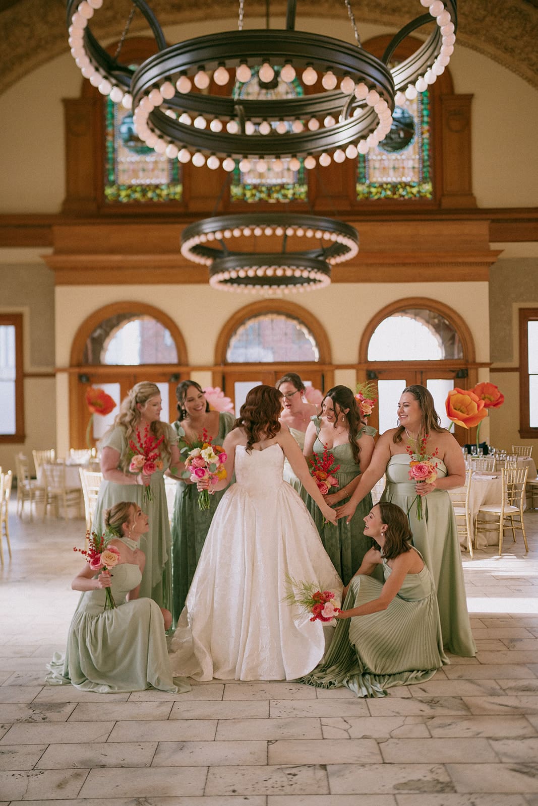 A bride in a white gown stands surrounded by seven bridesmaids in sage green dresses, all holding bouquets, inside a large, elegant hall with chandeliers at The Ashton Depot in Fort Worth