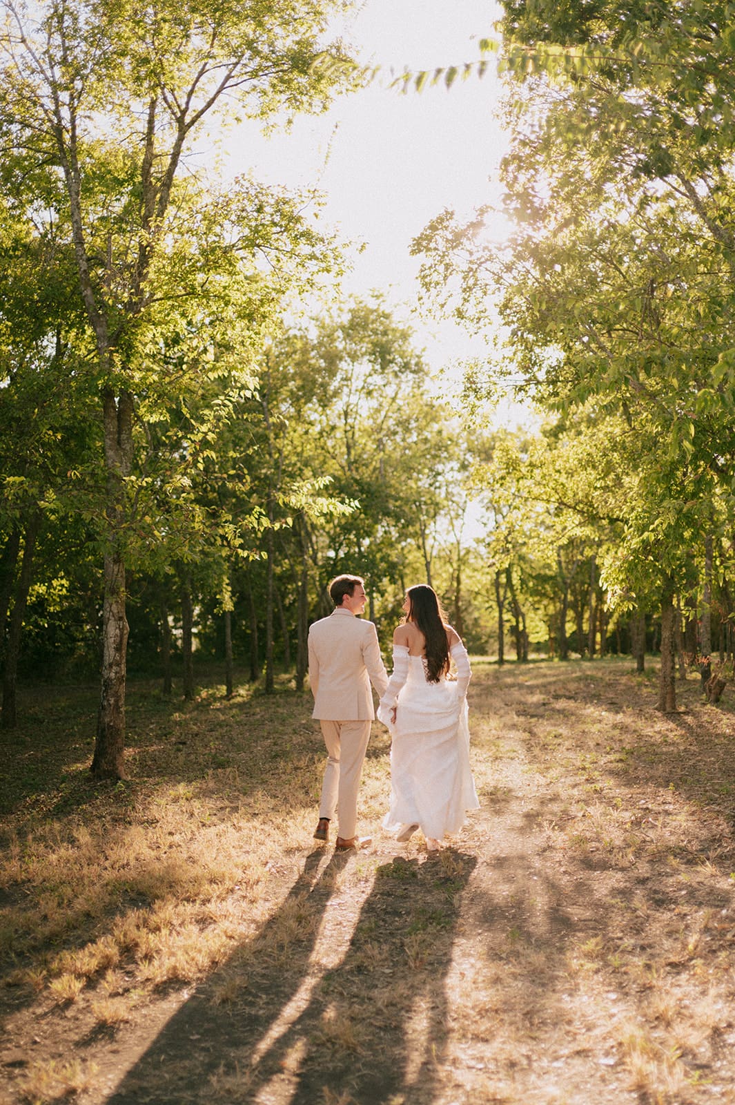 A bride and groom stand outdoors facing each other, holding hands in sunlight, with the bride holding a large bouquet of pink flowers for a backyard wedding in texas