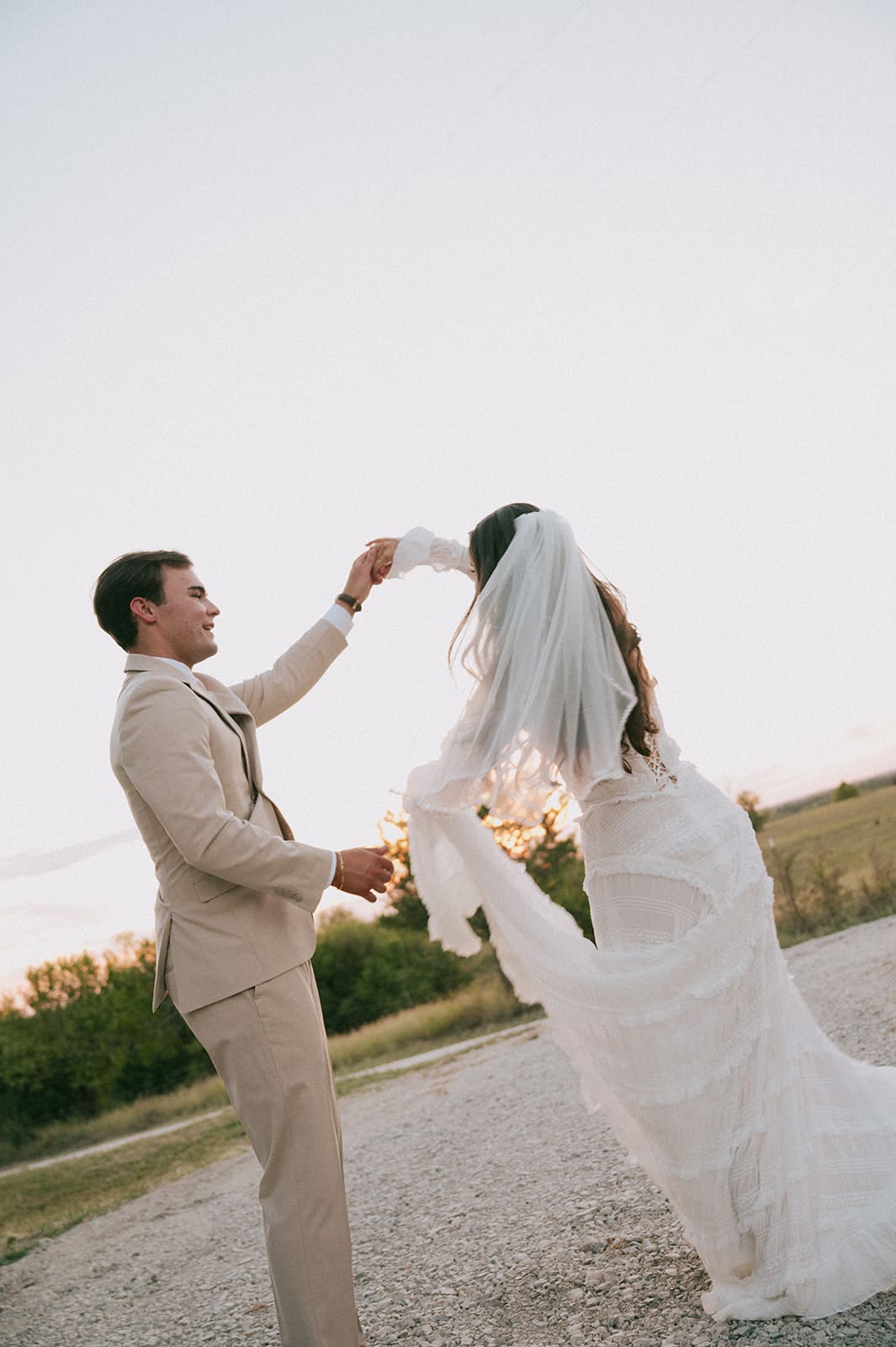 A bride in a white dress and veil stands beside a groom in a beige suit outdoors on a gravel path, with trees and fields in the background for a backyard wedding in texas