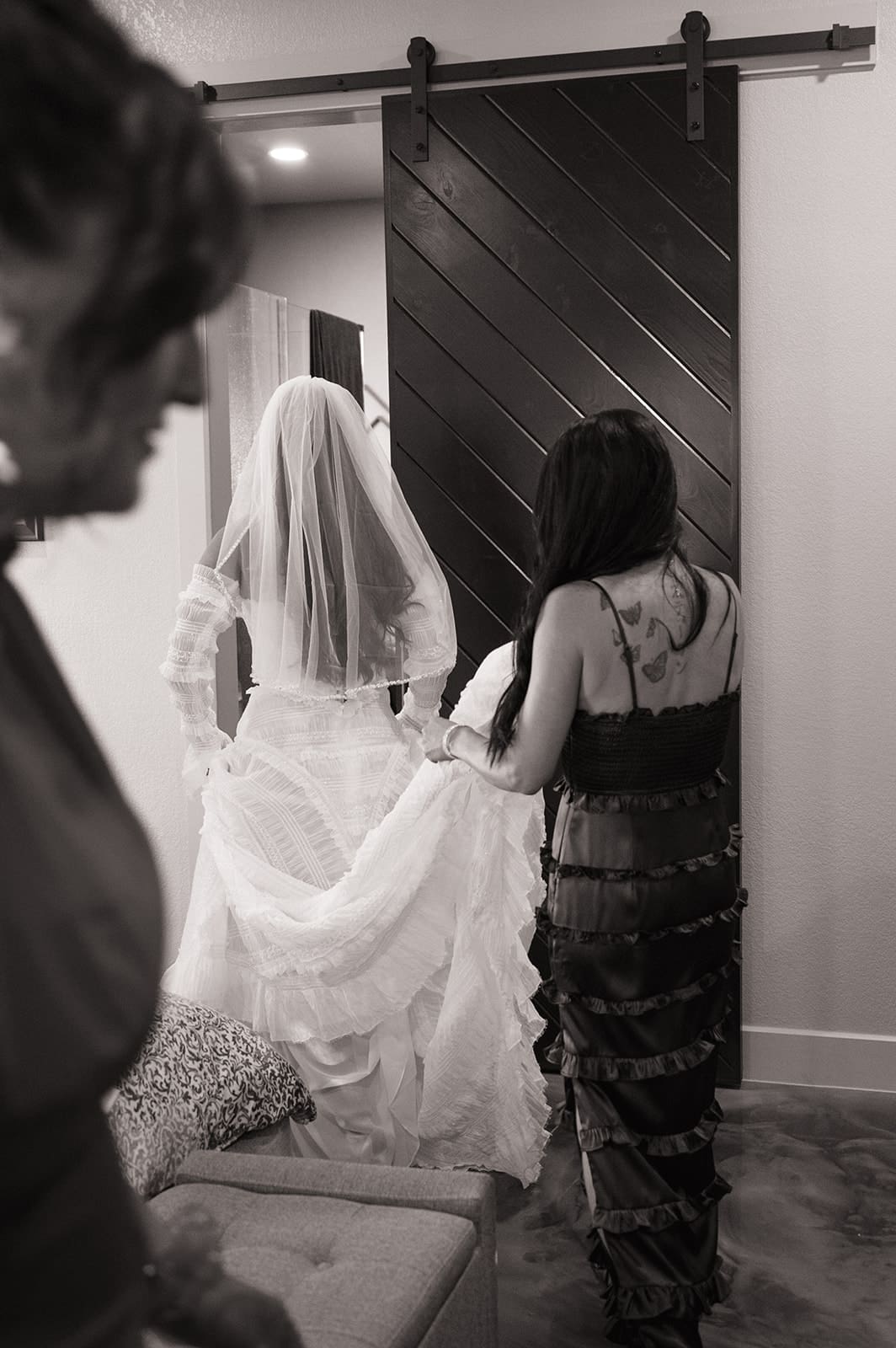 A bride in a wedding dress and veil walks with a woman adjusting her train; another woman stands in the foreground. The scene is indoors, in black and white.