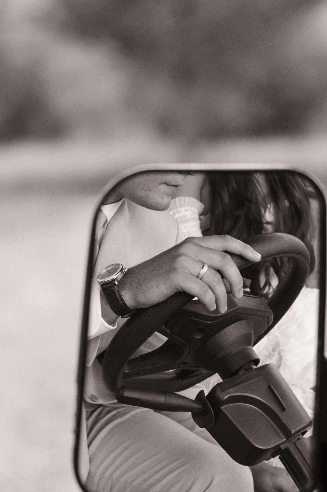 Close-up of a person's hand on a steering wheel, reflected in a side mirror, with part of their face and upper body visible.