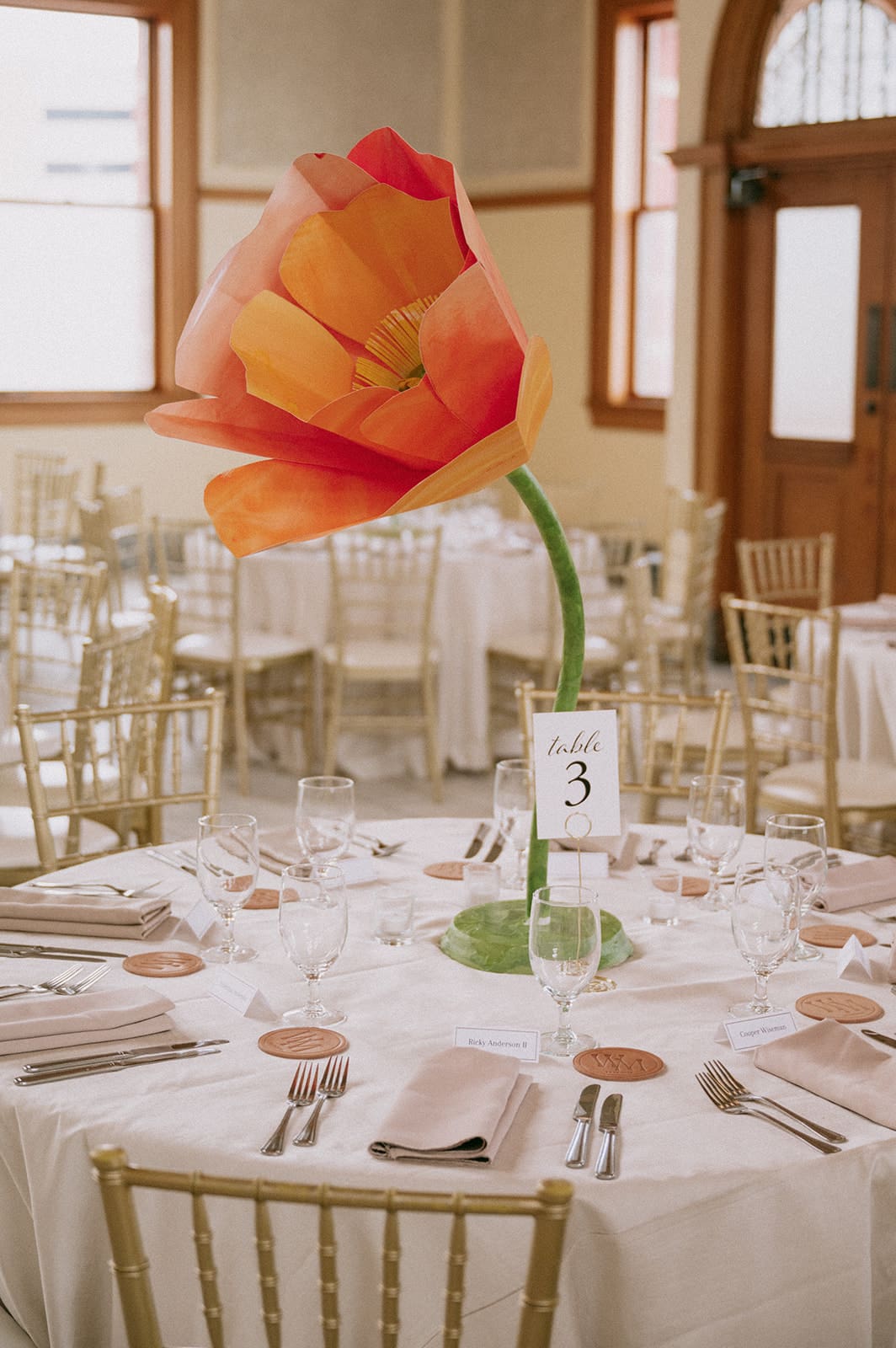 Elegant banquet hall with round tables, white chairs, oversized pink flower centerpieces, ornate stained-glass windows, and large circular chandeliers for a wedding at at The Ashton Depot in Fort Worth