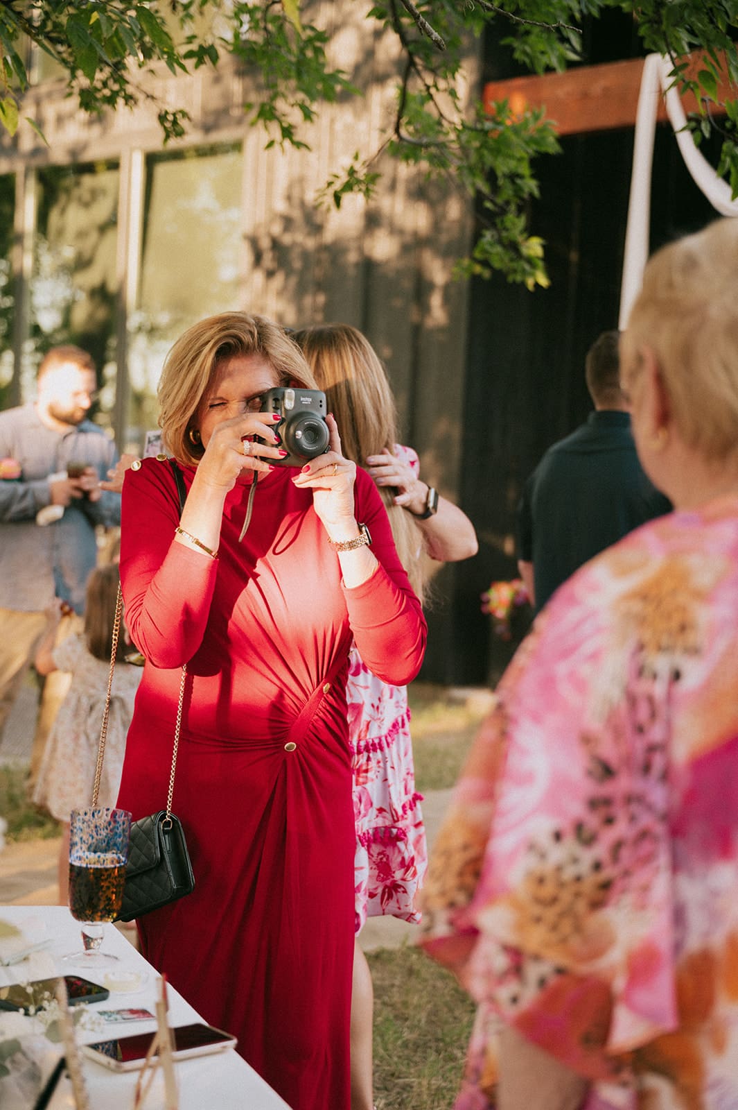 A woman in a red dress takes a photo with a camera at an outdoor event, with other people mingling in the background.