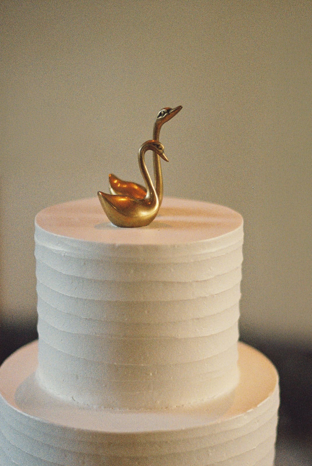 A white, two-tiered cake with horizontal textured lines, topped with a gold swan figurine.