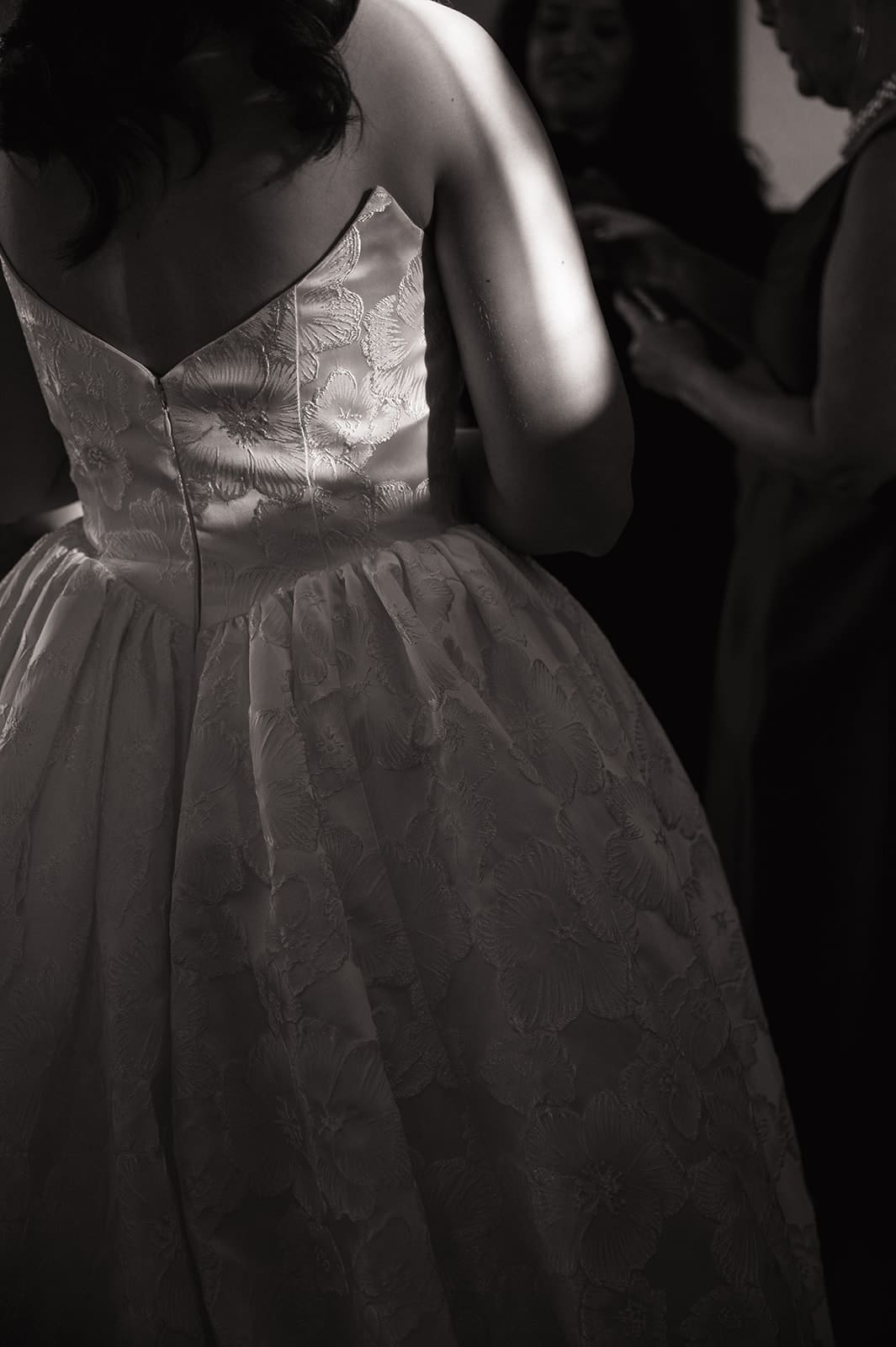 A person in a sleeveless, textured dress stands in the foreground with a group of people in dim lighting in the background for a wedding The Ashton Depot in Fort Worth