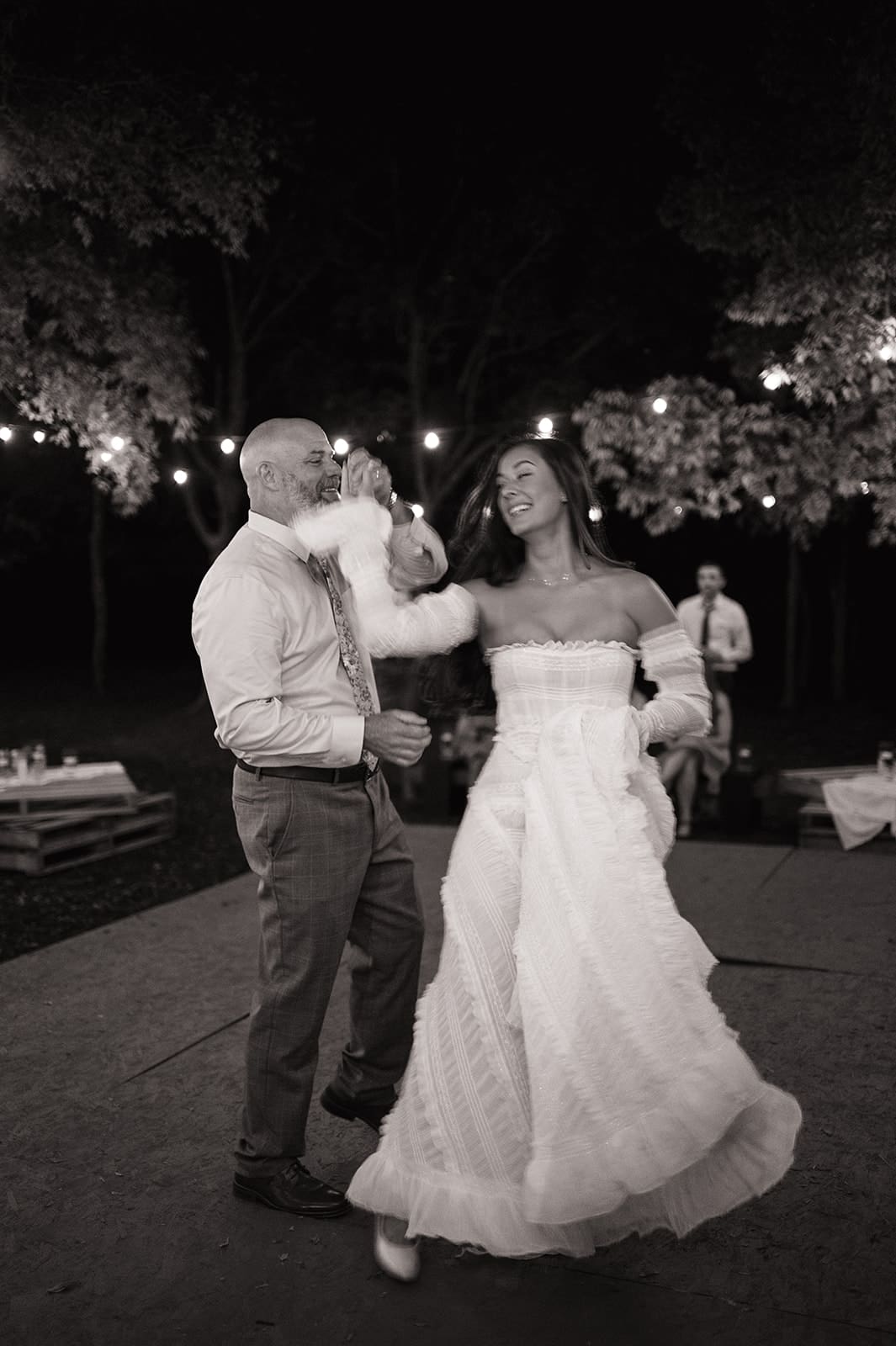 A bride in a white dress and her dad in a beige suit dance together outdoors at night under string lights for a backyard wedding in Texas