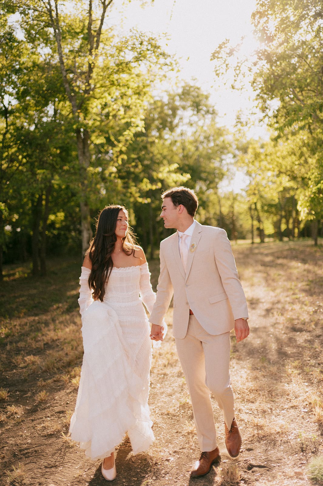 A bride and groom stand outdoors facing each other, holding hands in sunlight, with the bride holding a large bouquet of pink flowers for a backyard wedding in Texas